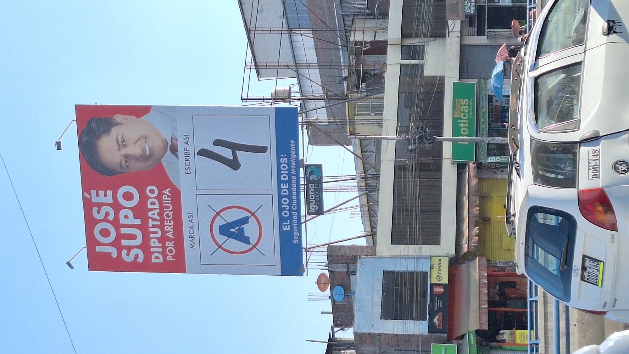 Here's a detailed description of the image:
**Scene:** The image appears to be taken on a street in José Luis Bustamante y Rivero, Peru. A political campaign billboard is prominent.
**Subjects:**
* **José Supo:** A political candidate for "Diputado Por Arequipa." His portrait is on the billboard.
* The billboard promotes the number "4" for the election, along with the text "Escribe Asi" (Write like this).
**Objects:**
* **Billboard:** A large political campaign billboard featuring José Supo.
* **Cars:** White cars are parked on the street. The license plate of one car is visible with the registration "DOD-148".
* **Buildings:** Several buildings are in the background, including commercial establishments. There are signs for a pharmacy "Boticas Perfumeria," and a store named "Tinka".
* **Overhead Wires:** A network of wires is visible, indicating electrical or communication lines.
* **Street Infrastructure:** A street sign, and a satellite dish on one of the buildings.
**Environment:**
* **Urban Setting:** The environment is a typical urban street in Peru.
* **Daylight:** The image was taken during the daytime.
**Overall Impression:** The image captures a street scene dominated by a political campaign sign.
