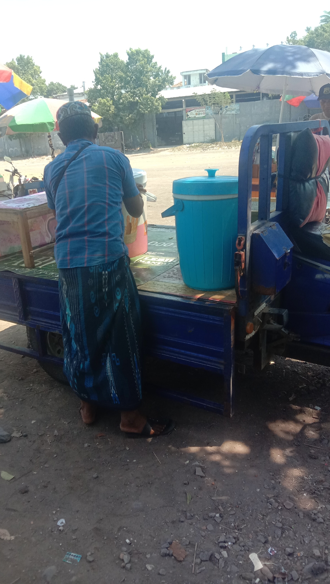 A person, viewed from behind, stands beside a blue truck in what appears to be the city of Geneng Waru, Indonesia. The individual is wearing a blue checkered shirt and a sarong with a dark blue and black pattern. They are holding a pink and white jug. To their right, a large blue cooler sits on the truck bed, and behind that, a dark-colored seat with a pink item on it is visible. The truck has a blue metal frame and bed. In the background, there are trees, a building with a white roof and gray walls, and umbrellas. The ground is a mixture of dirt and gravel. The lighting suggests it is daytime.