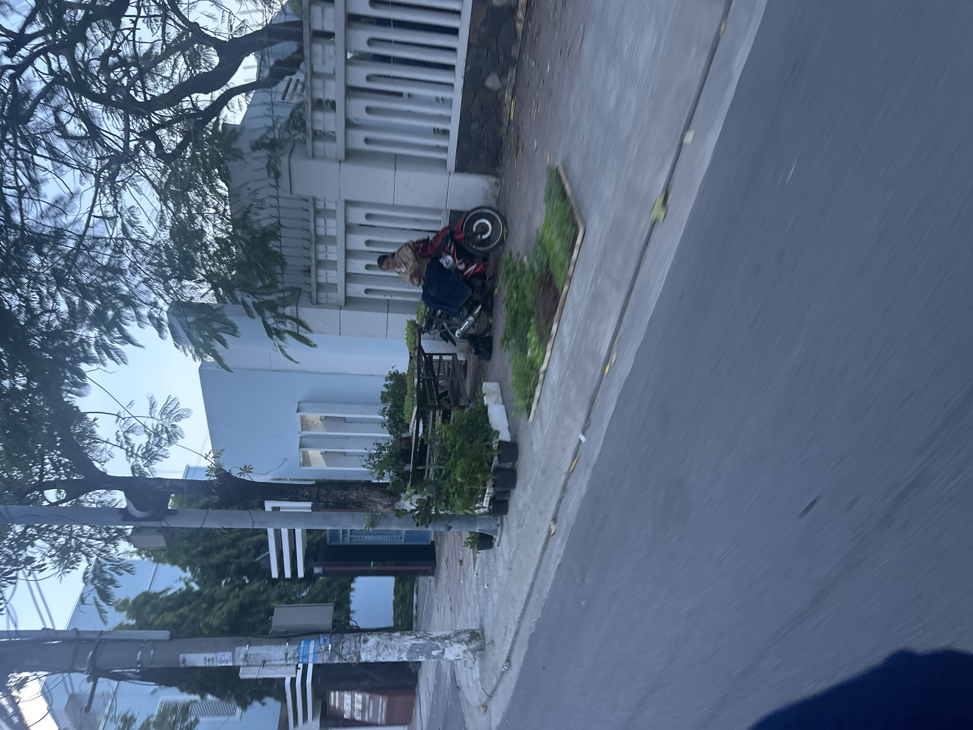 The image depicts a street scene in Bac Nha Trang Ward, Vietnam. A person is seated on a red and silver motorcycle parked on a sidewalk next to a white building with a gated facade. The sidewalk abuts a road. Trees and vegetation are visible along the sidewalk. A utility pole stands near the lower portion of the image.