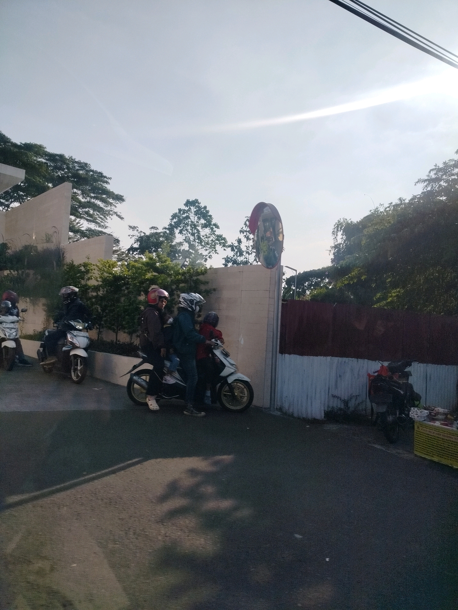 In Cisarua, Indonesia, a group of people on motorcycles are gathered near a wall and trees. The scene appears to be on a street or pathway.
In the foreground, the asphalt surface shows shadows cast by trees and possibly a line marking. To the left, a white motorcycle with a rider wearing a dark helmet is parked. Further to the left, another white motorcycle is visible, also with a rider.
In the middle ground, a white motorcycle carries three individuals. The rider wears a red helmet, and two passengers, one in the middle and one behind, are also on the motorcycle. To their left, another motorcycle with two riders, both wearing helmets, is positioned. The rider in front has a dark helmet, and the passenger behind has a red helmet.
The background features a light-colored, modern-looking wall with some greenery planted along its base. Behind the wall, lush green trees and foliage are abundant, suggesting a natural or well-maintained environment. On the right side of the image, a darker, corrugated metal fence runs along the property line. A convex security mirror is mounted on a pole next to the wall, reflecting the surroundings. Another motorcycle, darker in color, is parked near the fence on the right, with a stall displaying goods.
The sky is bright with a hazy sun, indicating it's daytime, likely morning or late afternoon due to the long shadows. The overall impression is a common street scene in a tropical or subtropical urban area, with motorcycles being the primary mode of transportation.
