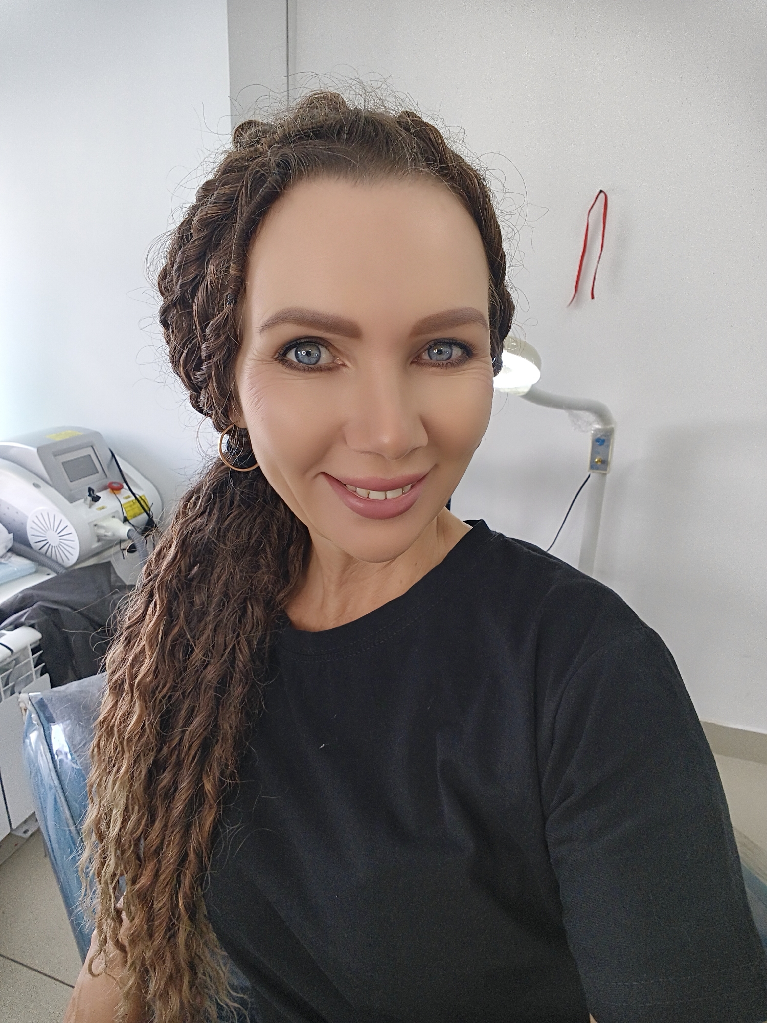 A woman with braided dark blonde hair and blue eyes smiles at the camera. She is wearing a black t-shirt and a hoop earring in her left ear. Behind her, a white wall is visible, with a red ribbon hanging from the ceiling on the right. To the left, a piece of medical equipment with a screen and a fan is on a table. A lamp with a white arm and a light bulb is also visible on the right, attached to the wall. The setting appears to be an indoor environment, possibly a medical or beauty clinic.