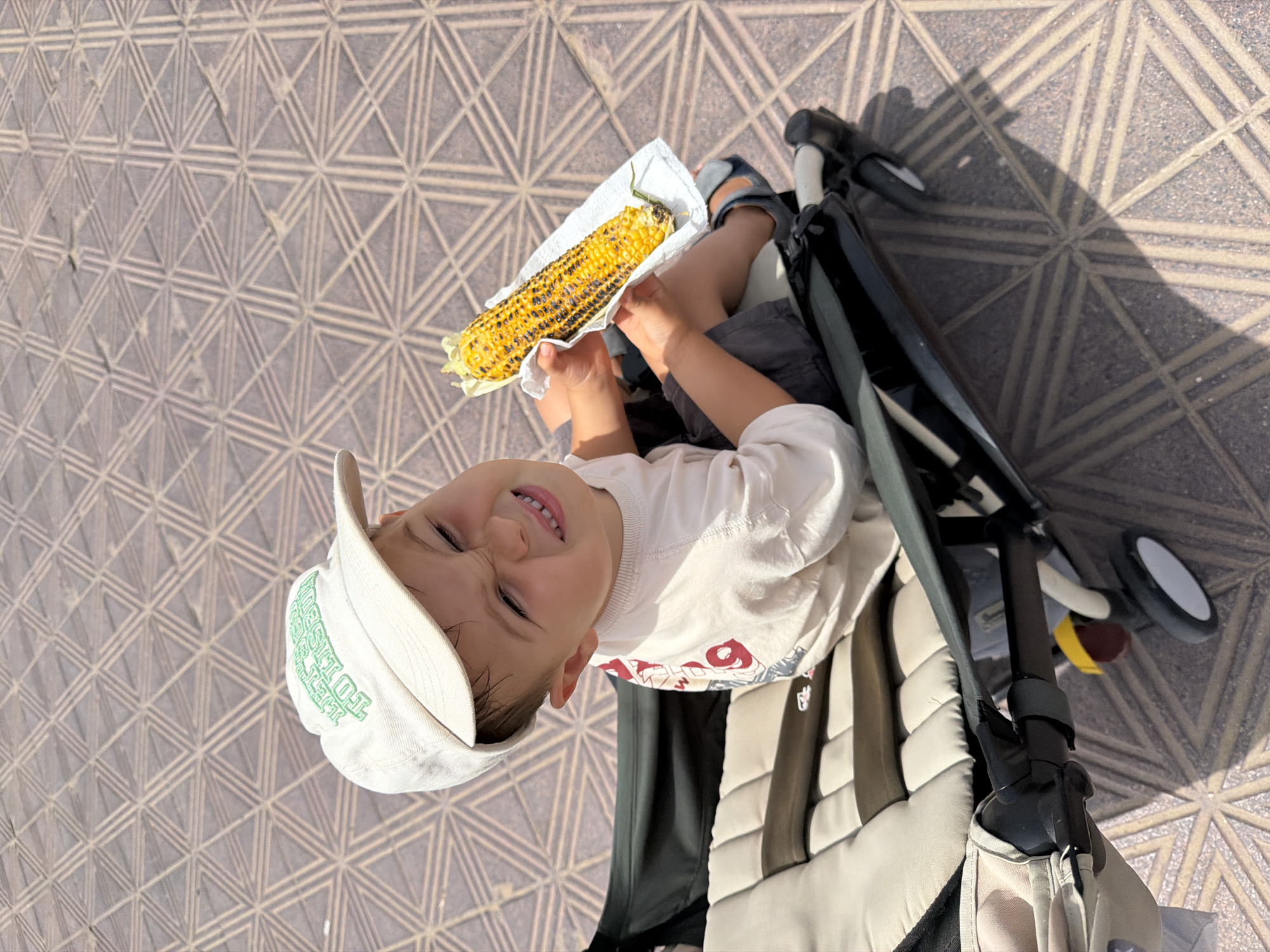 This is a candid, high-angle, outdoor shot of a young child sitting in a stroller, holding a piece of grilled corn. The child, wearing a white t-shirt and a white baseball cap, looks up and smiles at the camera. The corn is on a white paper napkin. The child's legs are visible, wearing dark shorts and sandals. The stroller has a dark frame and a beige and brown padded seat. The background is a patterned stone or tile ground, likely a sidewalk or patio, with geometric shapes. The lighting suggests it is daytime, possibly sunny, as there are shadows on the ground. The overall mood appears cheerful and casual, indicating a pleasant outing. The text on the child's cap is partially visible but unreadable. The text on the child's t-shirt is also unreadable.