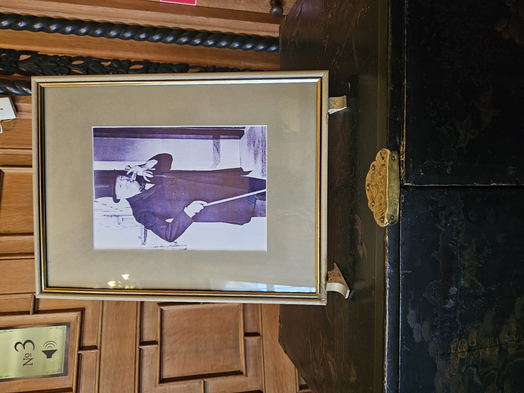 The image shows a framed black and white photograph mounted on a wooden wall, possibly inside a building. The photograph depicts a man in a dark coat and hat, holding a baton or pointer. The photograph is framed with a gold and tan frame. To the right of the framed picture is a dark, glossy cabinet or door, with decorative gold hinges and escutcheon. Below the framed picture, on the wall, there is a plaque that reads "No 3" next to a speaker icon. The wall and objects appear to be located in Алупкинский городской совет, Ukraine, based on geographical indications. The overall environment suggests a historical or formal setting.
