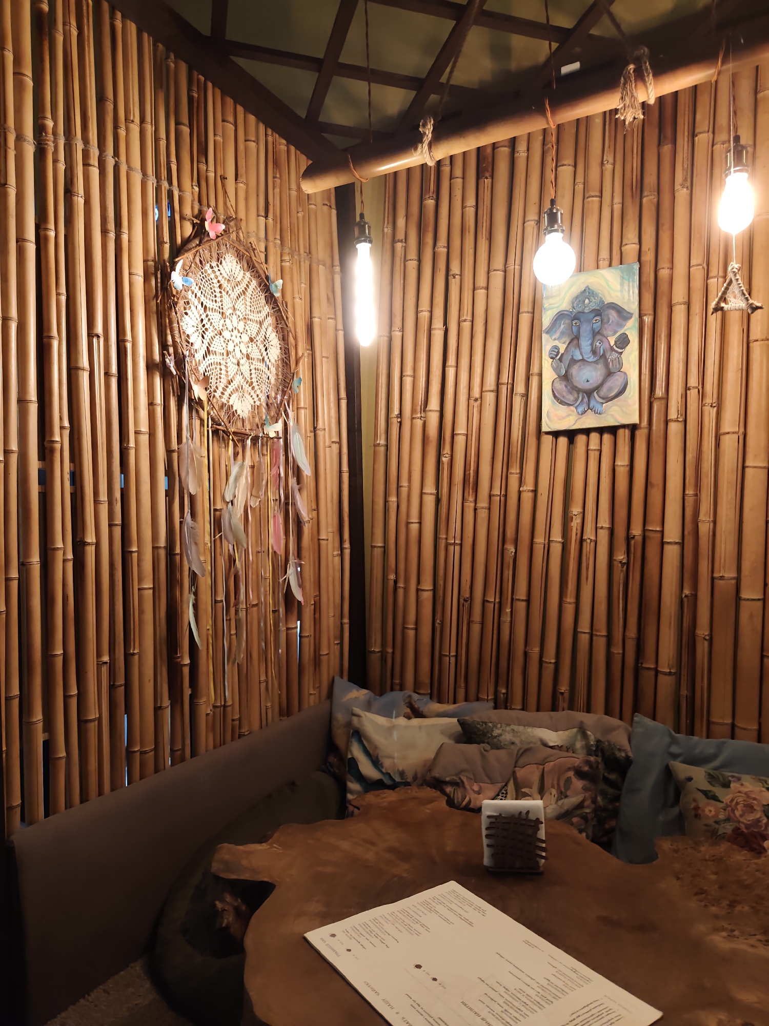 The image depicts the interior of a dimly lit cafe or restaurant with walls made of vertical bamboo poles. In the left half of the image, a dreamcatcher with feathers and small butterfly figures hangs on the bamboo wall. Above the bamboo, a wooden lattice ceiling is visible. Two Edison-style lightbulbs hang from the ceiling by cords, illuminating the space. To the right of the dreamcatcher and below one of the lightbulbs, a small painting of the Hindu deity Ganesha is hung on the bamboo wall. A comfortable seating area with cushioned benches and various patterned pillows is arranged along the wall. In the foreground, a rustic wooden table with an irregular shape holds a menu or document and a small decorative item. The overall ambiance suggests a relaxed and bohemian atmosphere. The location is identified as a cafe in Sochi, Russia.