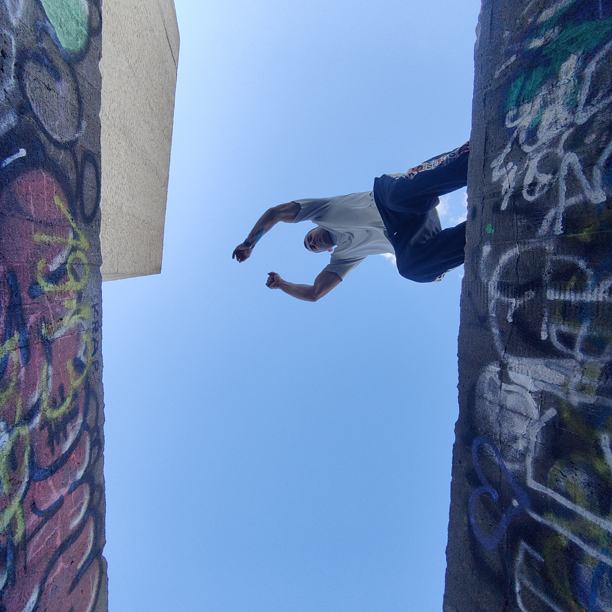 A low-angle shot captures a man performing parkour between two concrete structures covered in graffiti. The man is positioned horizontally against a clear blue sky. He wears a light-colored t-shirt and dark pants. The graffiti on the left structure features red, yellow, and green paint. The graffiti on the right structure is predominantly white. A building with a plain, light-colored exterior is visible in the upper left corner. The location is identified as Varna, Bulgaria.