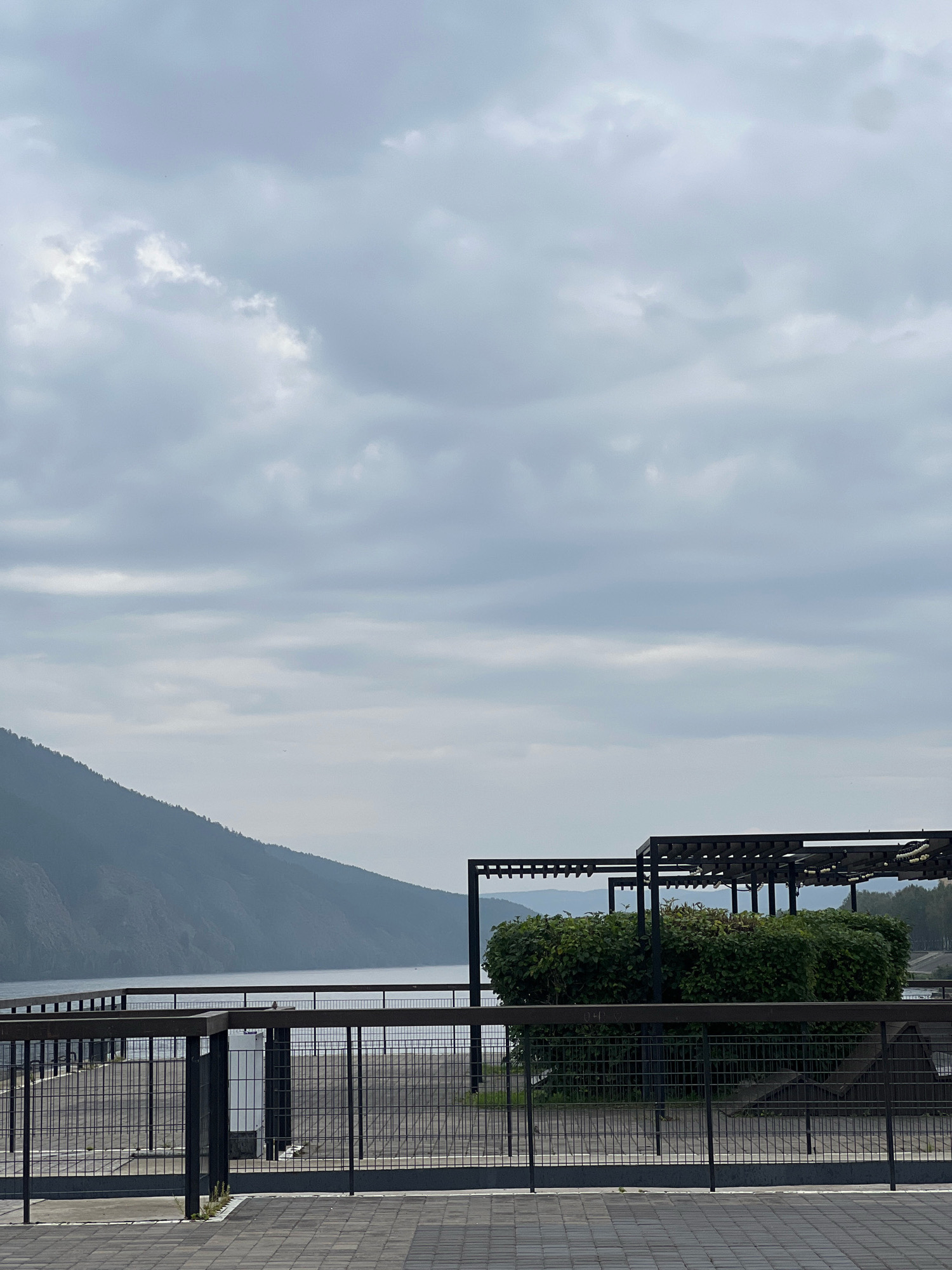 The image shows a cloudy day at a waterfront promenade in Divnogorsk, Russia. In the foreground, a paved area leads to a black metal fence and railing system. Beyond the fence, there is a walkway or plaza with a pattern of paving stones. Further back, a row of neatly trimmed green bushes is situated under a series of dark wooden pergolas. The water of a lake or river is visible behind the bushes and pergolas, with a large, forested hill rising from the opposite shore. The sky is overcast with various shades of gray clouds. The scene is quiet and appears to be unoccupied by people.