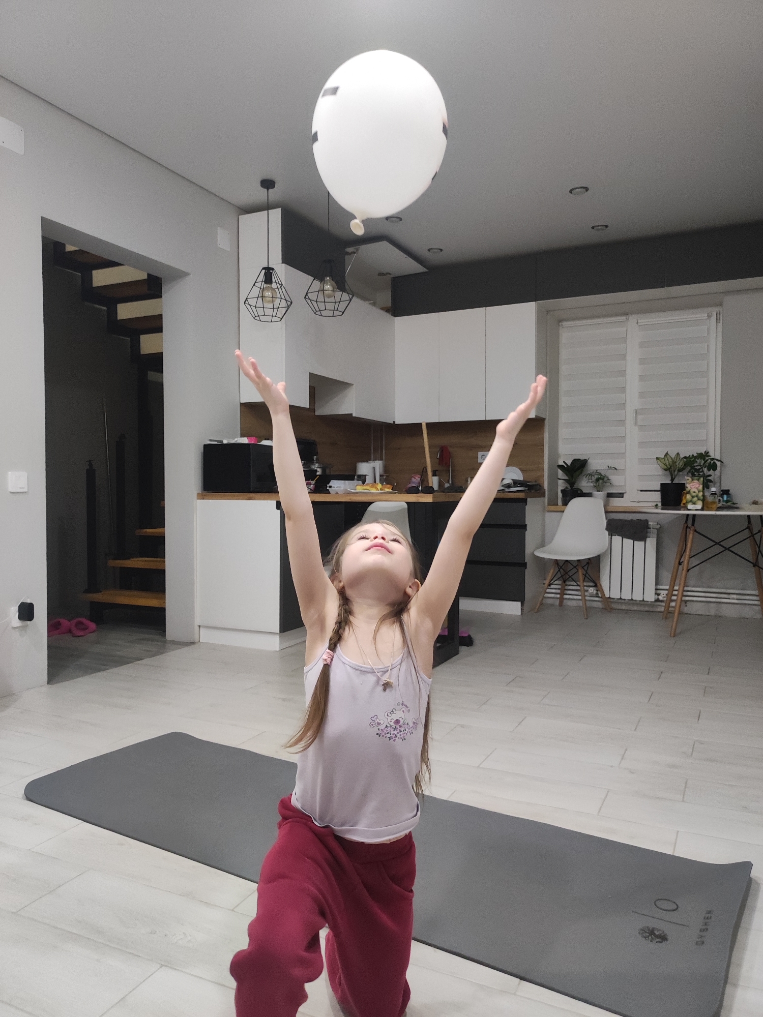 A young girl is indoors, likely in a home or apartment, standing on a gray yoga mat with both arms raised, seemingly attempting to touch or keep a white balloon aloft. The girl has long brown hair in a braid, a light purple top, and maroon sweatpants. The room appears to be a living and kitchen area, with a staircase in the background. The kitchen has white cabinets, black countertops, modern pendant lighting, and a window with blinds. There is a white chair and a table with plants near the window. The floor is light-colored. The image does not contain location information. Therefore, I am unable to provide an accurate location within Votkinsk, Russia.