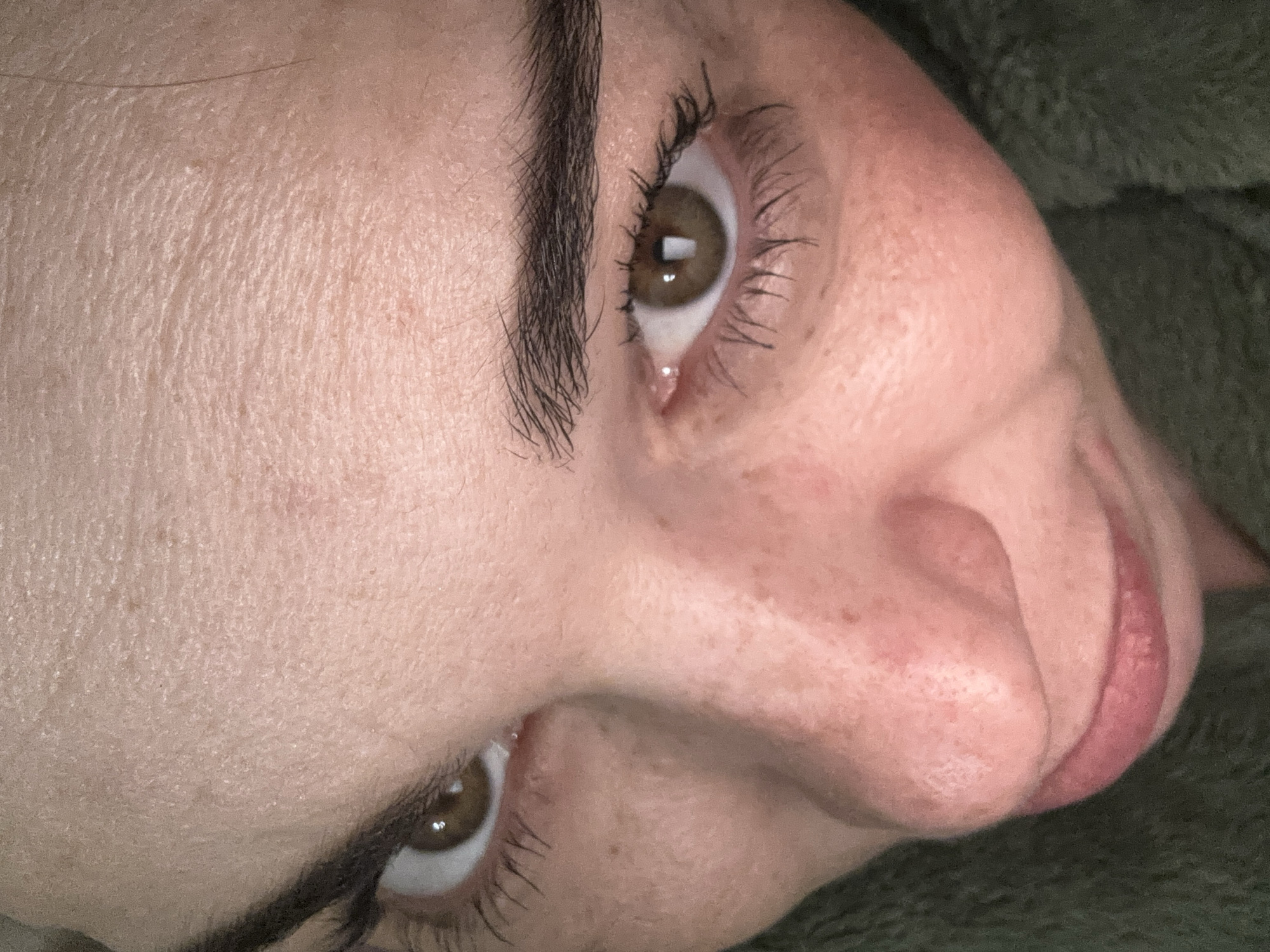A close-up, tilted view of a Caucasian female's face. Her complexion appears fair with visible pores and freckles on the nose and cheek. She has dark eyebrows and brown eyes with mascara on her upper eyelashes. Part of her nose and lips are visible in the foreground. The background is blurred and appears to be a green, textured fabric. No information indicates this image was taken in Vsevolozhsk, Russia.