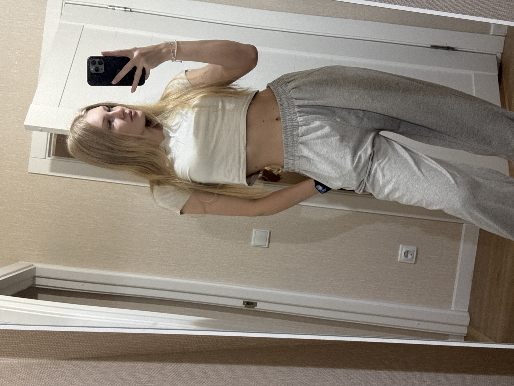 A young woman with blonde hair is taking a mirror selfie in what appears to be a residential hallway. She is wearing a light-colored crop top and grey sweatpants. Her left hand is in her pocket, and she is holding a black smartphone in her right hand to capture the photo. In the background, a beige textured wallpaper is visible, along with a white electrical outlet on the wall and a portion of a white door. The floor is made of light-colored wood. The overall impression is a casual, everyday scene within a home. The location is unknown, and there is no indication it is in the city of Perm, Russia.