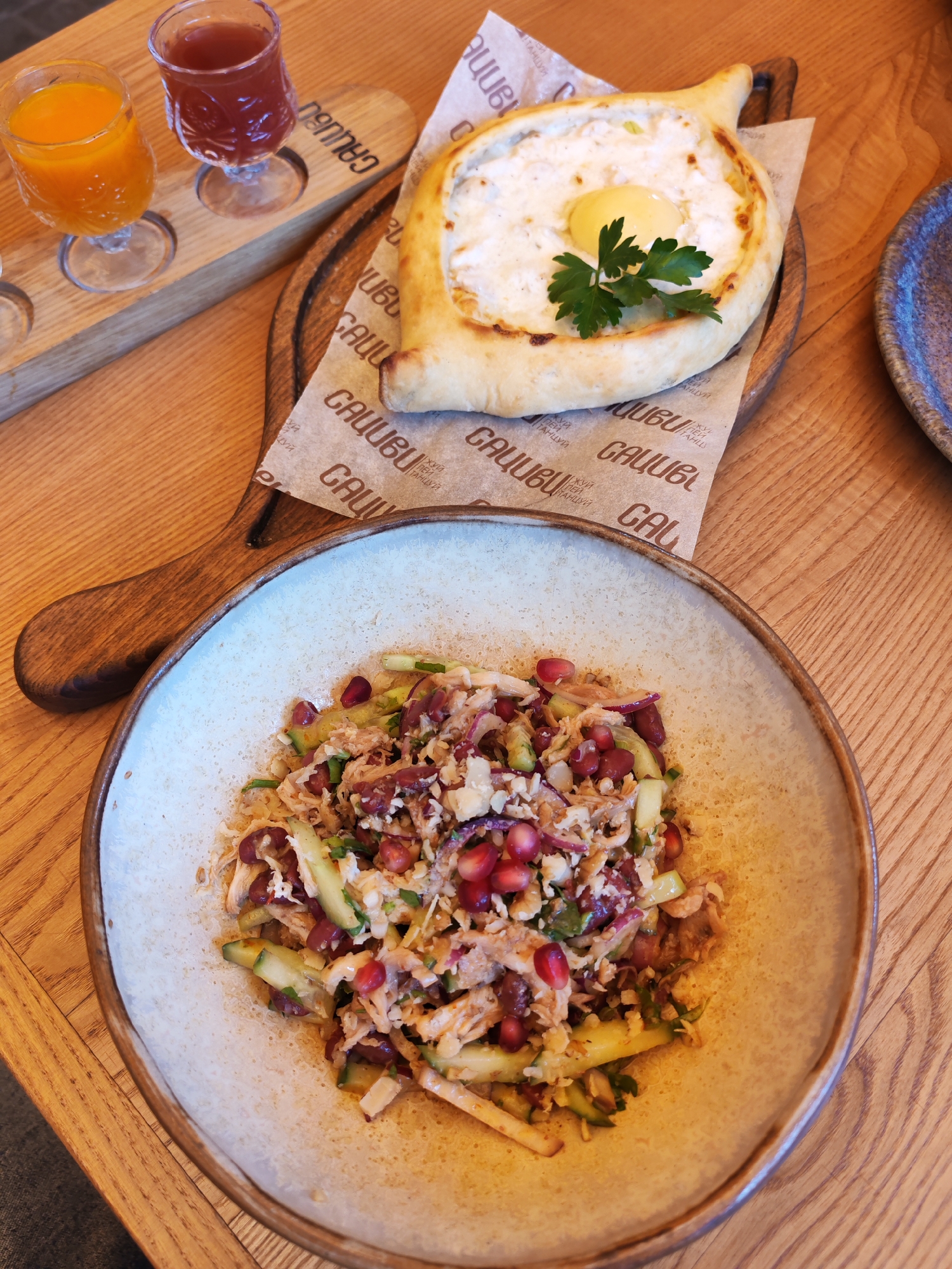 An overhead shot shows a table setting at a restaurant. In the foreground, a bowl contains a colorful salad with shredded chicken, cucumber, and pomegranate seeds. Behind the salad, a wooden board holds three small glasses filled with orange juice, a red beverage, and an empty glass. A portion of khachapuri, a Georgian cheese-filled bread with an egg in the center, rests on a piece of patterned paper on the wooden board. The table is made of wood, and a blue plate is visible on the right side of the frame. The restaurant appears to be located in Bol'shie Mkhi, Russia, as indicated by the restaurant's logo printed on the paper.