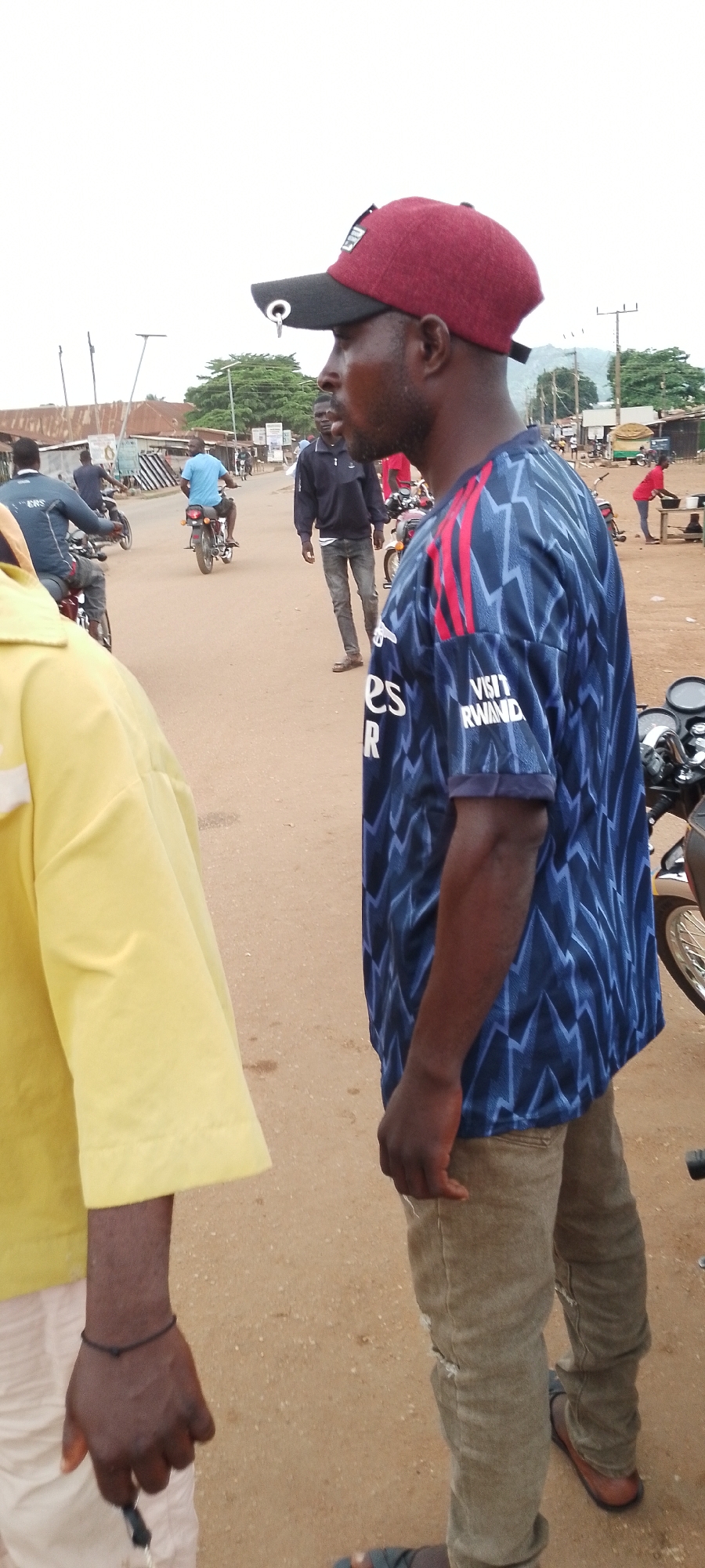 In Gboko, Nigeria, a man wearing a maroon and black baseball cap and a blue and red patterned shirt stands on a dirt road. The man is looking to his left, with his right hand on his hip. Behind him, several other people are visible, some riding motorcycles and others standing around. The environment appears to be a busy marketplace or street scene, with buildings and stalls lining the road. The sky is overcast, suggesting it may be daytime.