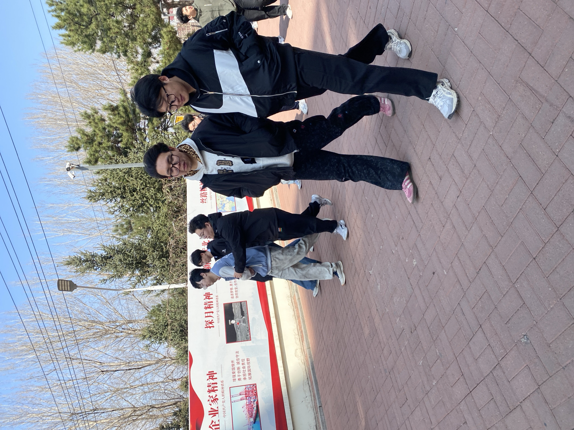 Here is a detailed description of the image:
The image appears to have been taken in an outdoor public area in the city of Zhongtun, China. A group of individuals are posing near a sign. The sign features Chinese text, including the phrase "企业家精神" (Entrepreneurial Spirit), "探月精神" (Lunar Exploration Spirit). The sign also displays images of a port and the moon.
In the foreground, two individuals stand close together, possibly posing or interacting. One of them wears a black jacket with a white stripe on the back and black pants, while the other wears a black and white hoodie with a leopard print hood lining and black pants. Both are wearing eyeglasses. Behind them, a group of other people is standing near the sign.
The background includes trees and bare branches against a bright blue sky, suggesting a time of year like late autumn or winter. A street light and some power lines are also visible. The ground appears to be a brick-paved walkway or plaza.