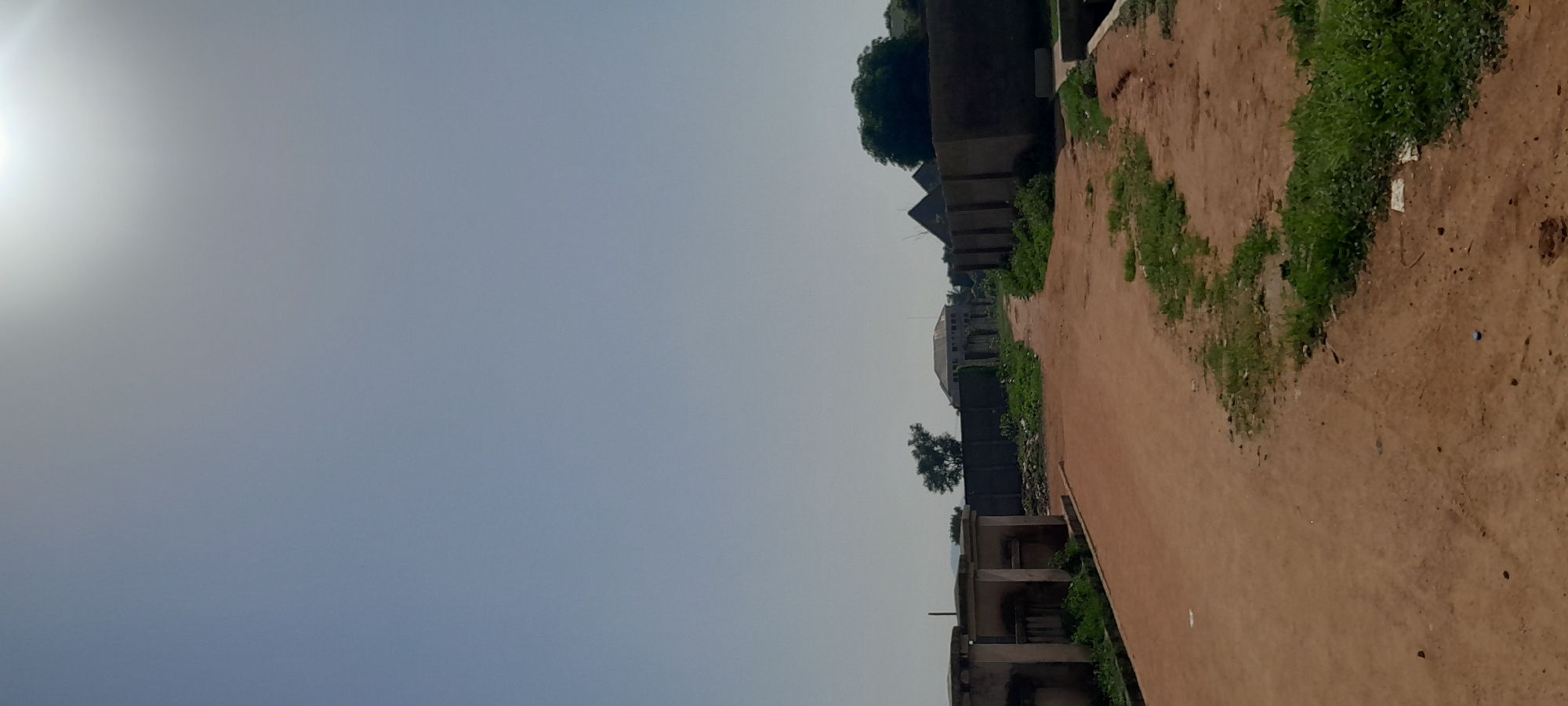 This media file depicts an outdoor scene in Jalingo, Nigeria. The image is oriented vertically, showing a clear sky on the left with the sun's glare indicating it is likely daytime, possibly morning or afternoon. A dirt path runs diagonally through the scene from the bottom right towards the top left.
Along the right side of the path are several structures. These appear to be unfinished buildings, characterized by exposed concrete and brickwork, with some of them featuring multiple levels and open doorways. Patches of green grass and vegetation are growing intermittently along the edge of the path and around the structures, suggesting a somewhat unkempt or developing environment. In the background, more substantial buildings are visible, including one with a domed roof, indicating a more developed area or a construction project of larger scale. A few trees are scattered throughout the scene, adding to the natural elements present.
There are no people visible in the image. The overall impression is one of a developing or partially constructed area in a warm climate, with the bright sun and earthy tones of the path being prominent features. The weather appears clear. No text is discernible in the image.