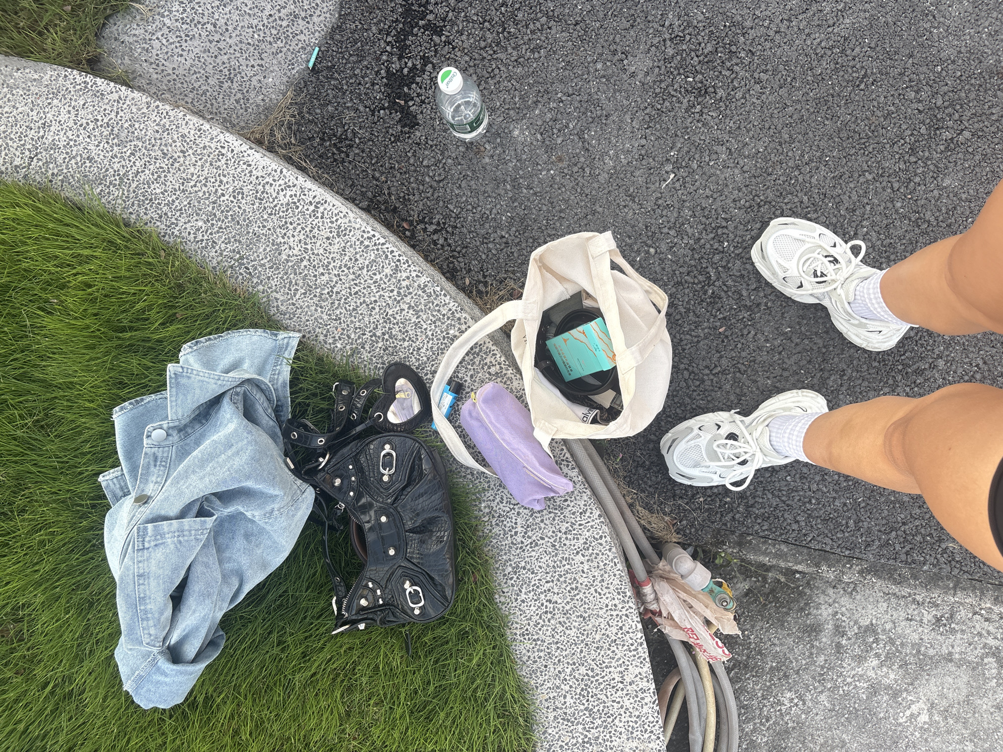 The image depicts a scene outdoors in what appears to be an urban park setting. The ground is composed of sections of green grass, grey speckled paving, and asphalt.
Objects present include a light blue denim jacket, a black studded purse, a purple pouch, a beige tote bag containing various items, a plastic water bottle, and several garden hoses.
A person is visible from the knees down, wearing white sneakers and socks. Their position suggests they are standing near the belongings scattered on the ground. The scene appears to be casually arranged, perhaps indicating a moment of rest or setting down belongings.
Without further information, determining the exact location in Haizhu District is not possible.