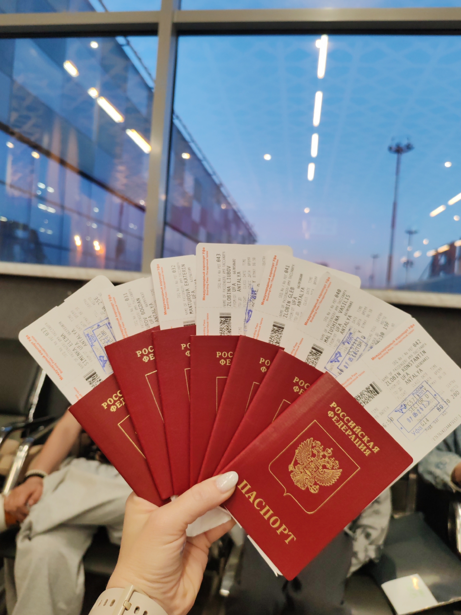 The image shows a hand holding five Russian passports and matching boarding passes. The boarding passes indicate flights from Ufa, Russia (UFA airport) to Antalya. The passports are red with gold lettering, indicating they are Russian Federation passports. A stamp on the boarding pass suggests a date of 07.06.2022. The background features a blurred view of what appears to be an airport terminal with large windows. The image was taken inside the Ufa International Airport in Ufa, Russia.