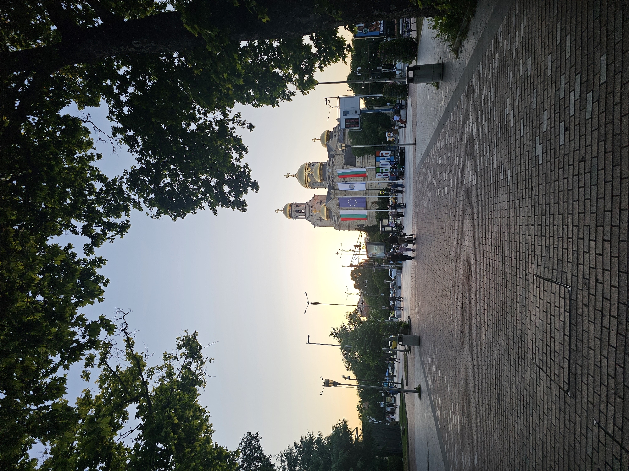 An outdoor, eye-level, long shot captures the Cathedral of the Assumption in Varna, Bulgaria, at dusk. The cathedral, a large, imposing structure with golden domes, is adorned with flags. In the foreground, a brick-paved walkway leads toward the cathedral, with trees framing the top of the shot. Several people are walking on the walkway, some with strollers. Streetlights are visible, and the sky is a soft gradient of orange and blue, indicating the end of the day. The scene is peaceful, with an atmosphere of evening strolls and civic pride.
