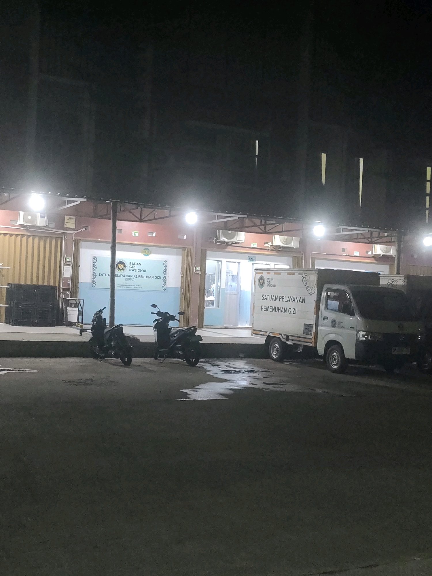 The image shows a nighttime view of a building with the text "BADAN GIZI NASIONAL SATUAN PELAYANAN PEMENUHIN GIZI" visible on a banner and on a white box truck. Two motorcycles are parked in front of the building. The building appears to be a commercial or government facility. The scene is illuminated by artificial lighting. There is no indication of the specific location within Batam, Indonesia, beyond the text on the banner and truck.