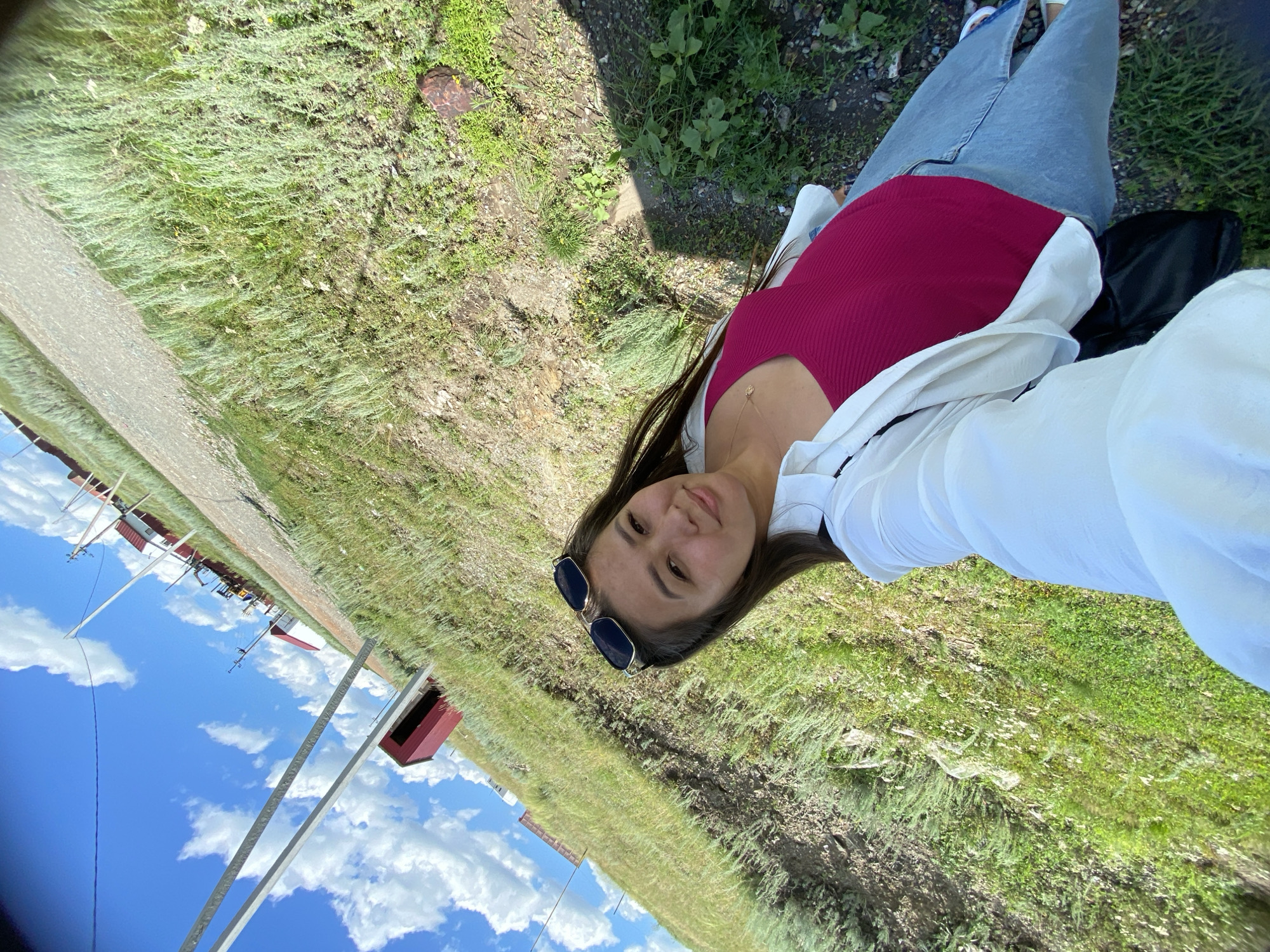 A young woman is taking a selfie outdoors on a sunny day. She is wearing a white button-down shirt over a magenta tank top, light blue jeans, and sunglasses on her head. She is standing on a grassy area next to a gravel path. In the background, there are several red and white buildings and utility poles against a bright blue sky with white clouds. The scene appears to be in a rural or semi-rural setting.