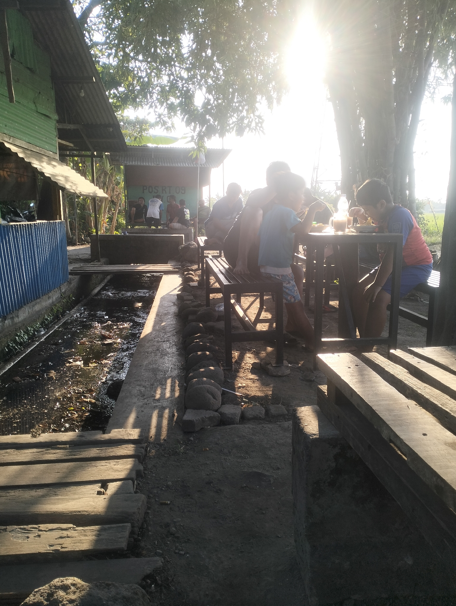 The image depicts a scene in Sambungrejo, Indonesia, with a group of people, primarily children, gathered around a table outside. The setting appears to be a rural or semi-urban environment, characterized by simple structures, a canal with murky water, and abundant trees. The sun is shining brightly, creating strong contrasts and lens flare.
In the foreground, a wooden walkway made of several planks is visible, leading towards the left side of the image. Adjacent to the walkway, to the left, is a green, corrugated metal building with a metal roof, possibly a house or a small shop. A blue corrugated metal fence runs along the front of this building. A narrow canal flows alongside the walkway, containing debris and dark water.
Further back, a low concrete structure with the sign "POS RT 09" is present. Several individuals are seated on benches or standing near this structure. A group of children and young adults are seated at a dark-colored metal table, some of them engaged in eating or drinking. The table is set with plates of food and drinks, including a clear bottle with a white cap and a glass or mug.
The people in the image are mostly silhouetted or partially obscured due to the strong backlighting from the sun. The children at the table are wearing casual clothing. The environment is somewhat unkempt, with evidence of natural growth and some litter in the canal. The overall impression is of a community gathering or a mealtime in a modest outdoor setting, illuminated by the bright Indonesian sun.