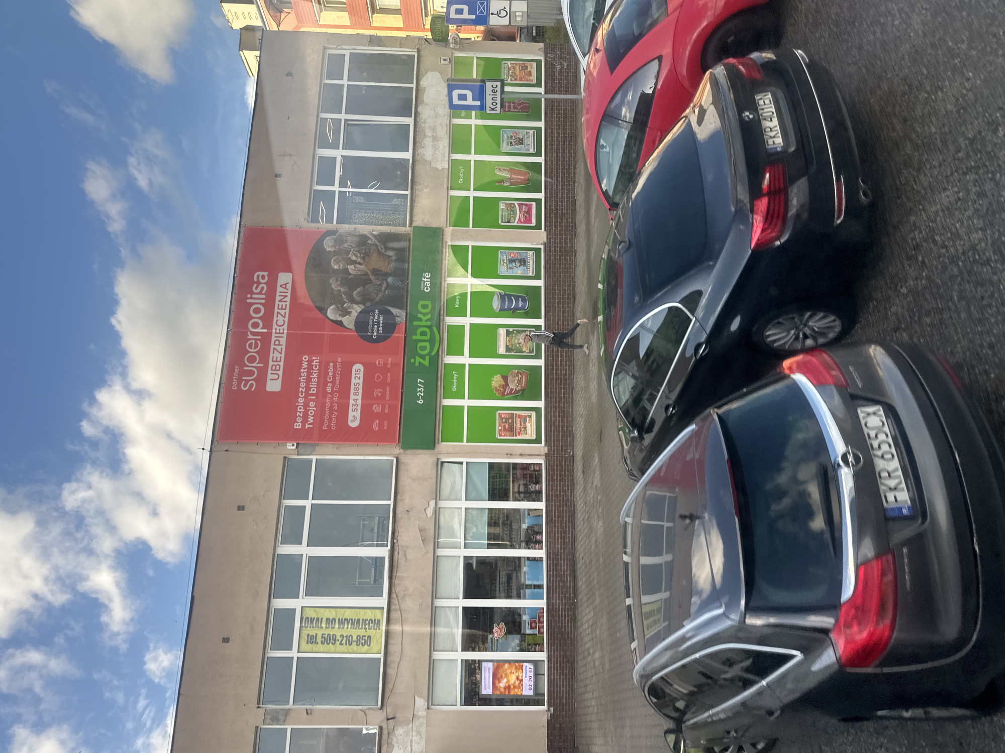 A street scene in Krosno Odrzańskie, Poland, features a modern commercial building with storefronts. A large red banner with white text advertising "superpolisa UBEZPIECZENIA" (insurance) dominates the upper portion of the building. Below this, green awnings mark the entrance to a "Żabka" convenience store. The store displays various promotions on its windows, including images of food items and a person. A person is seen walking on the sidewalk in front of the store. Several cars are parked in front of the building, including a dark gray Opel Insignia with the license plate "FKR 655CX" and a black BMW with the license plate "FKR 401EN". The sky above is partly cloudy.