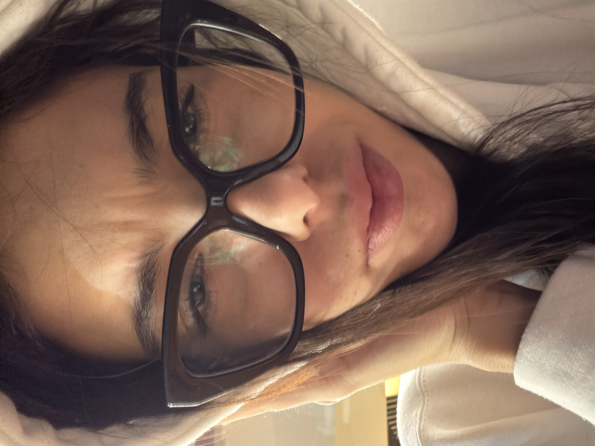 The image is a close-up selfie of a young woman wearing large, dark-rimmed glasses and a white hoodie. She has her right hand on her cheek and is looking directly at the camera with a slightly furrowed brow and a neutral expression. Her hair is dark and falls around her face. The lighting suggests an indoor setting, possibly illuminated by a soft, warm light source. There are no discernible architectural or environmental features that can be used to determine a specific location within Sofia, Bulgaria.