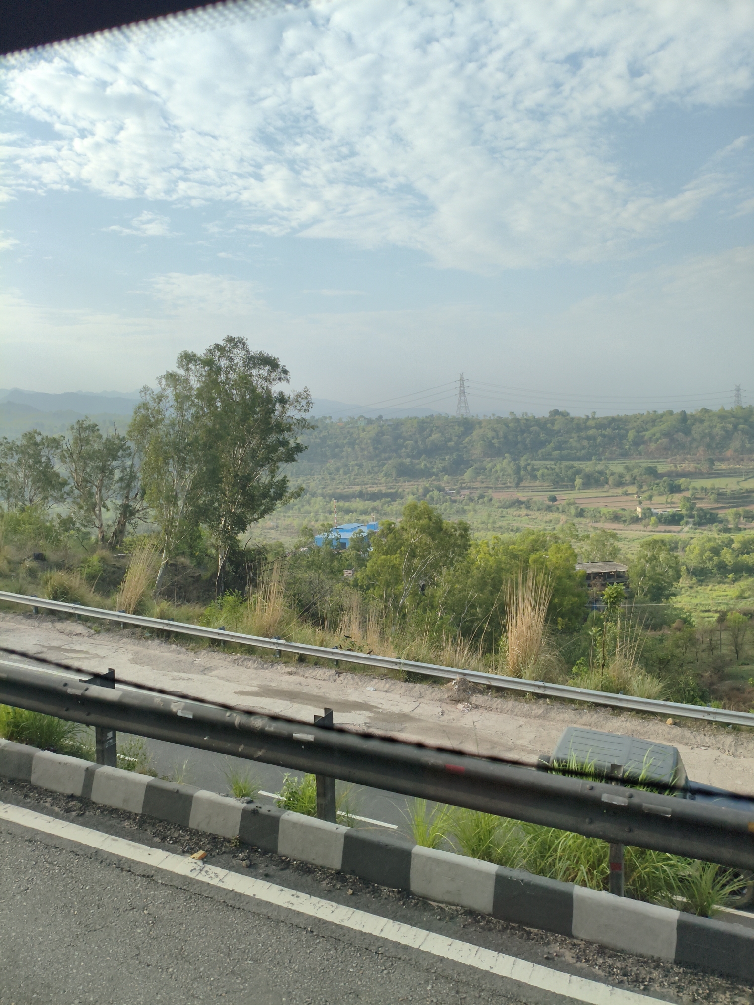 The image shows a view from a road overlooking a valley in Bilaspur, India. The foreground features a road with a concrete barrier and guardrail. Beyond the road, there is a lush green valley with rolling hills, dotted with trees and some scattered buildings. The sky is partly cloudy with a hazy atmosphere. Power lines and transmission towers are visible in the distance, indicating the presence of infrastructure. The overall scene suggests a scenic, rural or semi-rural environment.