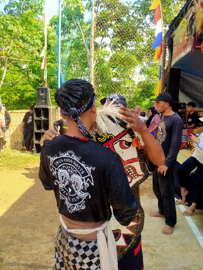 An outdoor scene in Plosorejo, Indonesia, depicts a cultural event under bright daylight. In the foreground, two male figures are central. The figure on the left is seen from behind, with dark, wet hair tied by a black and white bandana. He wears a black long-sleeved shirt with a white graphic on the back featuring the text "SINGO KRIS BUDOYO" and two stylized head profiles, along with a white sash over a black and white checkered sarong-like garment. His left arm is around the right shoulder of the adjacent figure. The figure on the right is partially visible, wearing a dark cap and holding a decorated, horse-shaped prop with red, white, and black patterns and white, brush-like ornamentation on its head. His right hand is raised with an open palm.
In the midground, to the right of the central figures, a male in a black long-sleeved shirt and cap stands barefoot, observing. Behind him, several individuals, including a woman in a pink top and head covering holding a child, are visible. To the far right, a makeshift structure with a blue tarp roof is adorned with red, white, and blue flags, and other decorative elements that resemble animal masks or heads. A young male is seated beneath this structure. To the left, a stack of large black speakers is positioned near a bamboo fence. Another male in a black shirt stands behind the speakers.
The background consists of dense green foliage and trees under a clear blue sky. Vertical poles with green netting are also visible. The ground in the immediate foreground is bare brown earth, transitioning to a lighter surface with a white painted line on the far right.