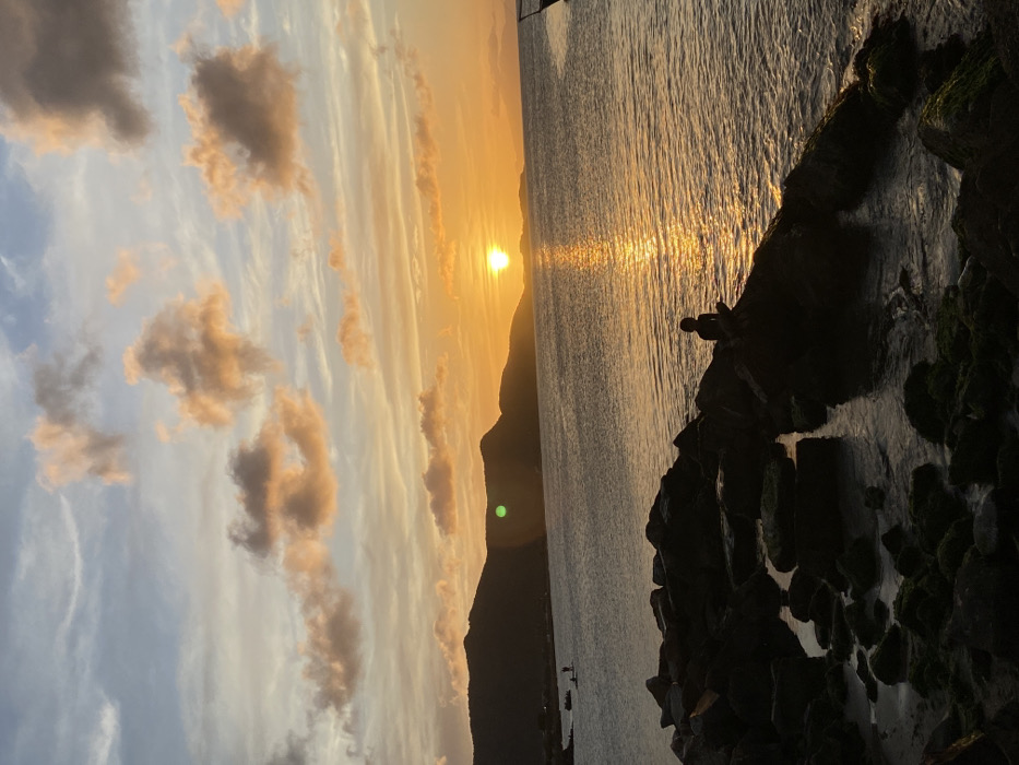 A breathtaking sunset scene unfolds with the sun's golden light reflecting across the water. Rocks line the shore, some covered with greenery. A lone silhouette sits upon the rocks. The sky is filled with scattered clouds, tinged with the warm colors of the setting sun. A mountain range is visible in the distance.