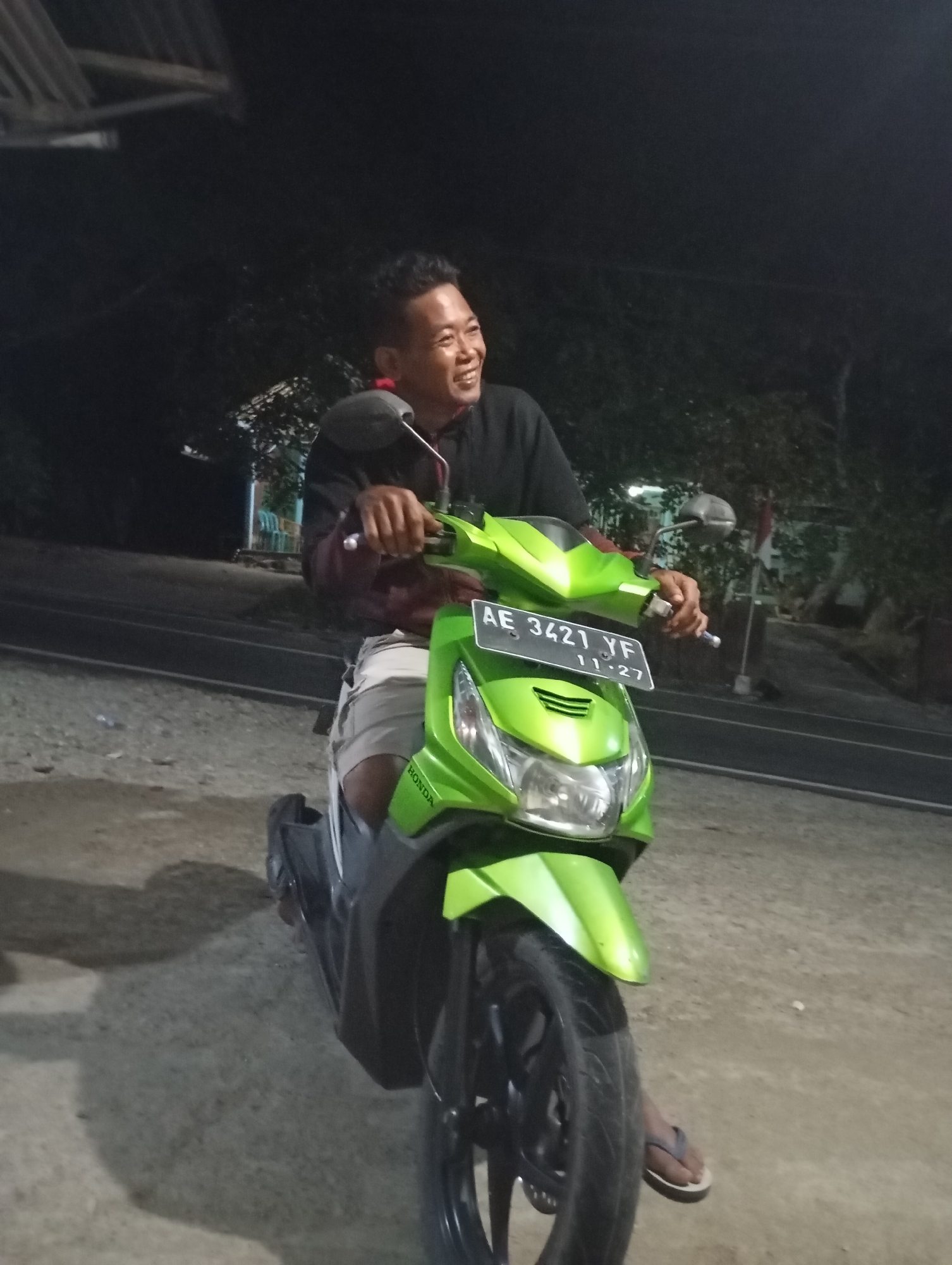 A man with short dark hair, wearing a black shirt and light-colored shorts, is sitting on a green motorcycle. He is smiling and looking to his right. The motorcycle has a license plate that reads "AE 3421 YF 11-27". The scene appears to be outdoors at night, with a dark background suggesting trees and possibly some buildings or structures. The ground is dirt or gravel. A shadow of the man and motorcycle is visible on the ground. The location is identified as Wonogondo, Indonesia.