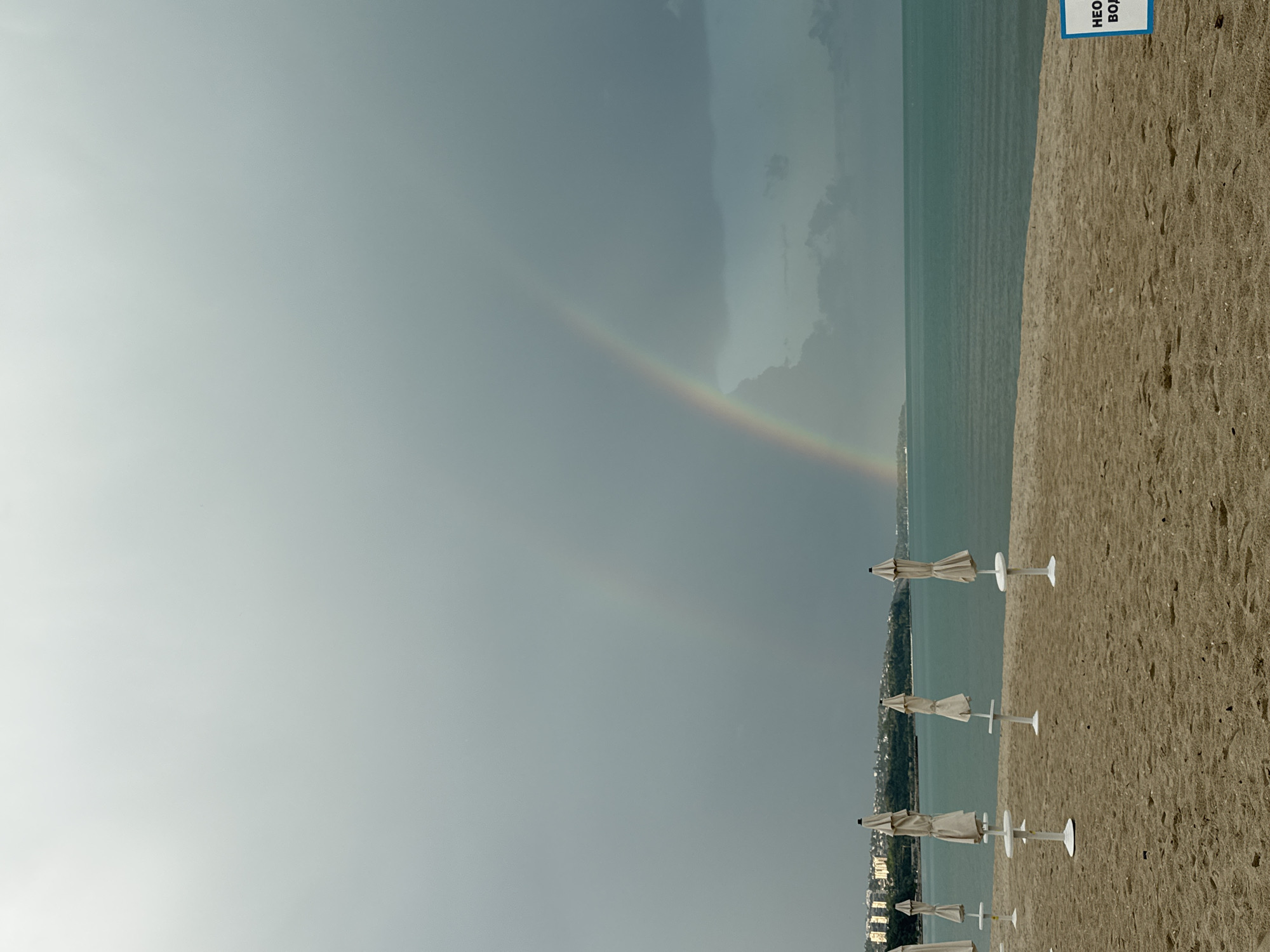 The image depicts a beach scene in Varna, Bulgaria. A subdued rainbow arcs across the sky. The beach is sandy and features a row of closed white beach umbrellas. The Black Sea is visible, meeting the horizon under a cloudy sky. The background shows a cityscape. The image is vertically oriented.