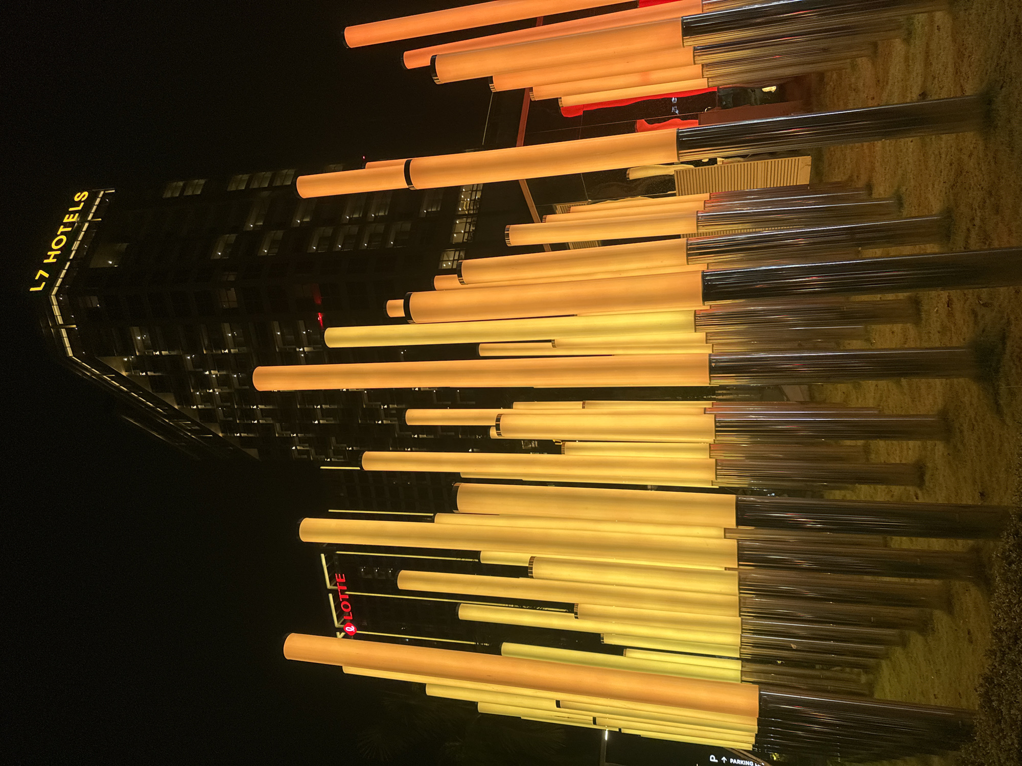 The image captures a nighttime scene in front of the L7 Hotels in Hanoi, Vietnam. A row of illuminated cylindrical pillars in varying shades of yellow and orange stands in the foreground, with their bases grounded in a patch of grass. In the background, the L7 Hotels building is visible, its facade illuminated with many small windows. Above the L7 Hotels sign, another sign reads "L7 HOTELS" in yellow lettering. To the left of the L7 Hotels building, a red "LOTTE" logo is also visible. The overall impression is of a modern urban landscape at night, featuring architectural lighting and decorative elements.