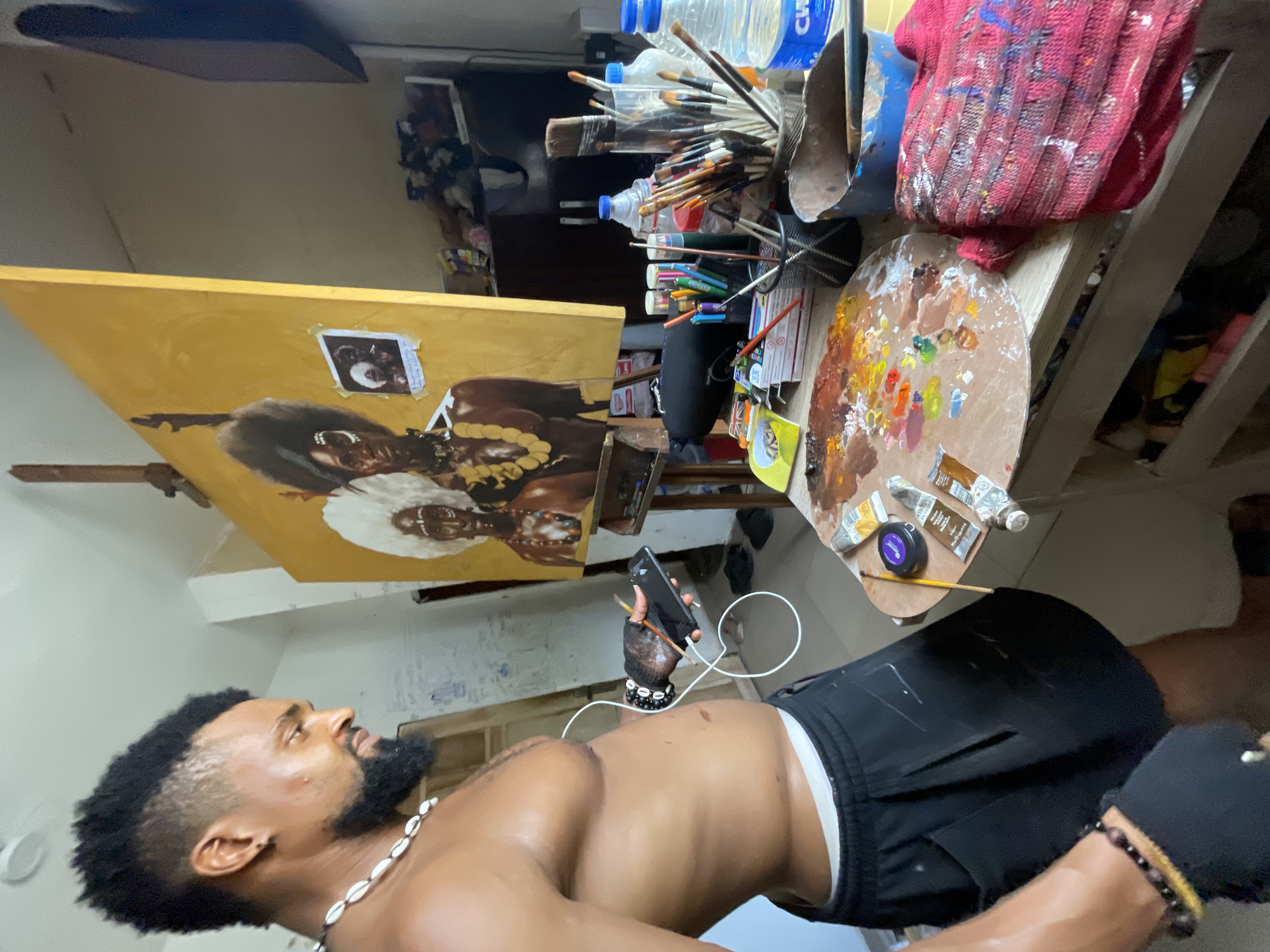 A man with dark skin, a shaved head with a thick afro on top, and a beard, is standing shirtless, wearing a white beaded necklace, black shorts, and a bracelet on his wrist, holding a smartphone in his left hand. He is in what appears to be an artist's studio. An easel holds a painting of two dark-skinned women with large afro hairstyles and necklaces. A small photograph is attached to the painting. To the right of the painting, there is a table with a variety of art supplies, including brushes, paint tubes, a paint palette, and water bottles. A red and blue paint-splattered cloth is also on the table. The background is a wall with some markings. Based on the prompt's instructions, the location is in the city of Ado, Nigeria, although there are no visual elements to confirm this.