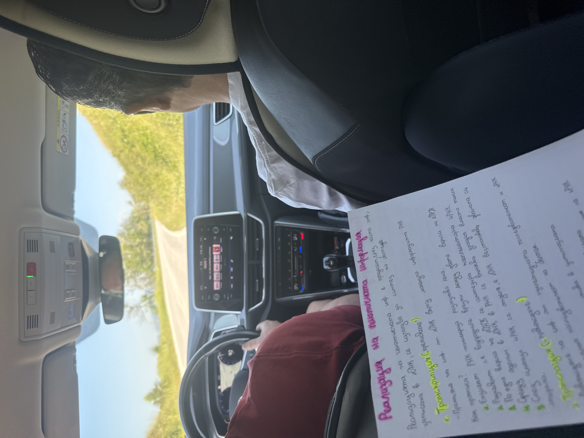 The scene depicts the interior of a modern vehicle in motion, viewed from the perspective of a rear passenger. Two individuals occupy the front seats. The driver, clad in a dark red long-sleeved shirt, is partially visible on the left, with a hand on the steering wheel. The front passenger, with short dark hair, is visible from behind, wearing a light-colored collared shirt, seated in a black leather seat with light-colored stitching. The dashboard features a digital display showing "92," and a central infotainment screen displaying "RADIO 1." An overhead console includes multiple buttons, a green indicator light, and a red SOS button. In the foreground, a white piece of paper with handwritten notes in Bulgarian Cyrillic script is prominently displayed. The notes, titled "Реализация на генетичната информация" (Realization of Genetic Information), detail biological processes such as transcription and translation, with subheadings highlighted in yellow. Outside the vehicle, a winding asphalt road traverses a landscape of green, vegetated hills under a clear blue sky. The environment appears rural and well-lit by daylight.