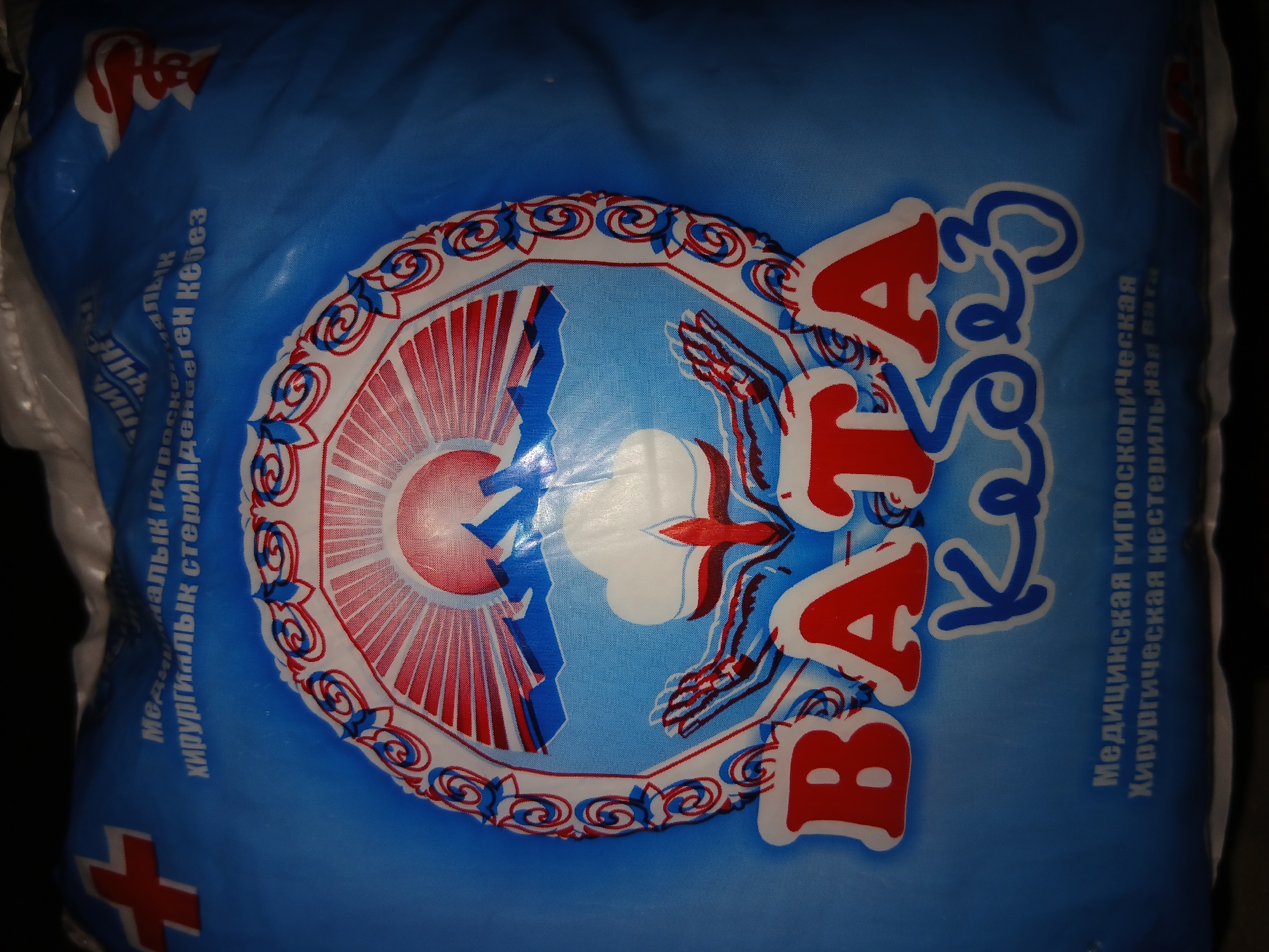 The image is a close-up, slightly blurry shot of a blue plastic bag, likely packaging for medical supplies. The bag features a prominent circular logo in the center with a sunrise over mountains, hands, and a cotton plant motif, all rendered in red, white, and blue. Large, bold red letters below the logo spell out "BATA" and smaller blue cursive letters below that read "Кебез". There is also text in Cyrillic script on the bag, including what appears to be "Медицинская гигроскопическая хирургическая нестерильная вата". A red cross symbol is visible on the upper left corner of the bag. The lighting is dim, creating a vignette effect around the edges of the image. The location in Тайгараев, Kyrgyzstan, is not visually depicted in the image itself.