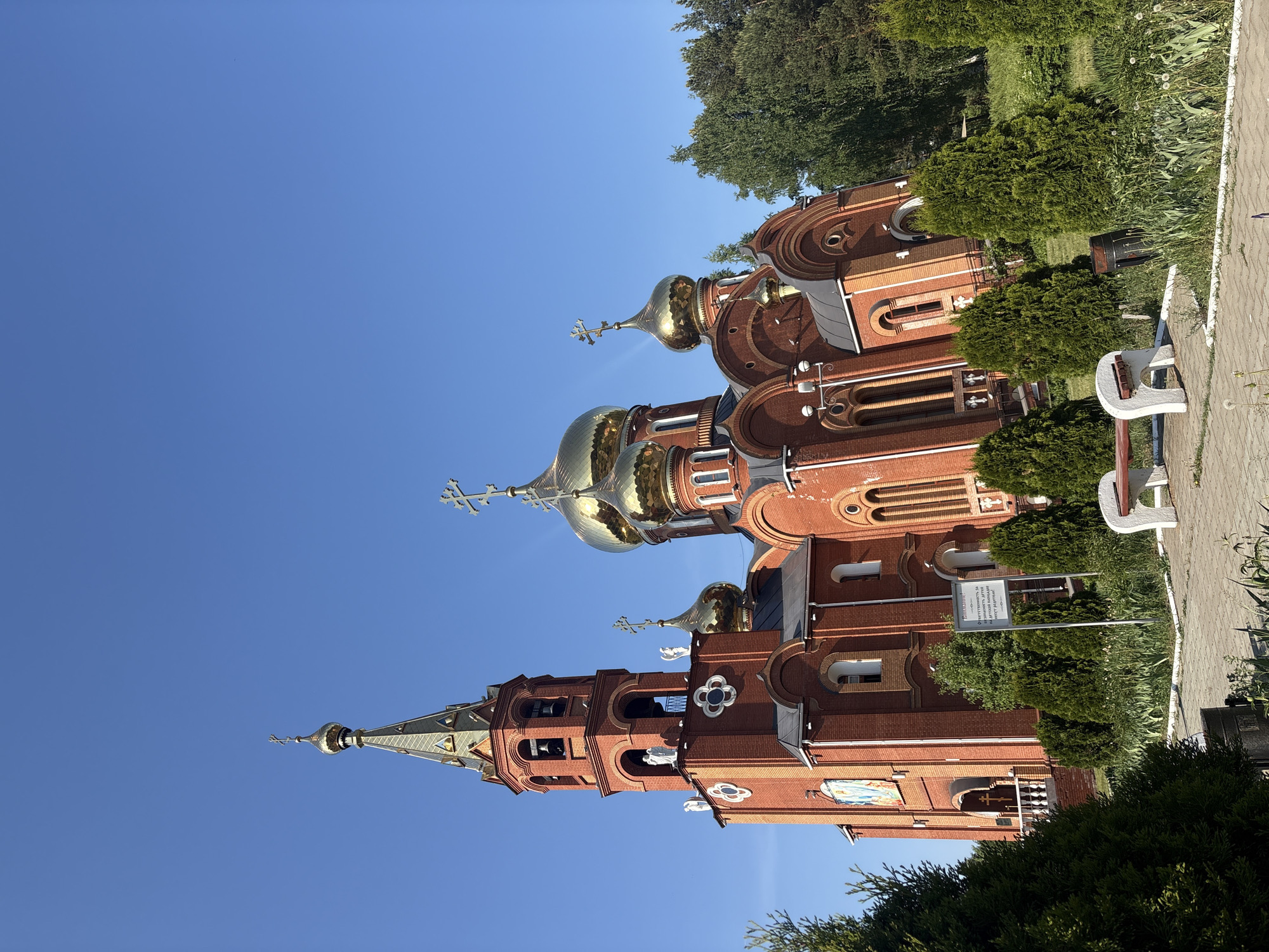 The image shows the Church of St. George the Victorious, located in Chaikovsky, Russia. The church is constructed of red brick and features golden domes topped with Orthodox crosses. Its architecture includes a tall bell tower and multiple arched windows. The sky is clear blue, suggesting a sunny day. The surrounding area includes green trees and bushes, and a paved walkway with a bench is visible on the right side of the image. A sign with Russian text stands near the church.