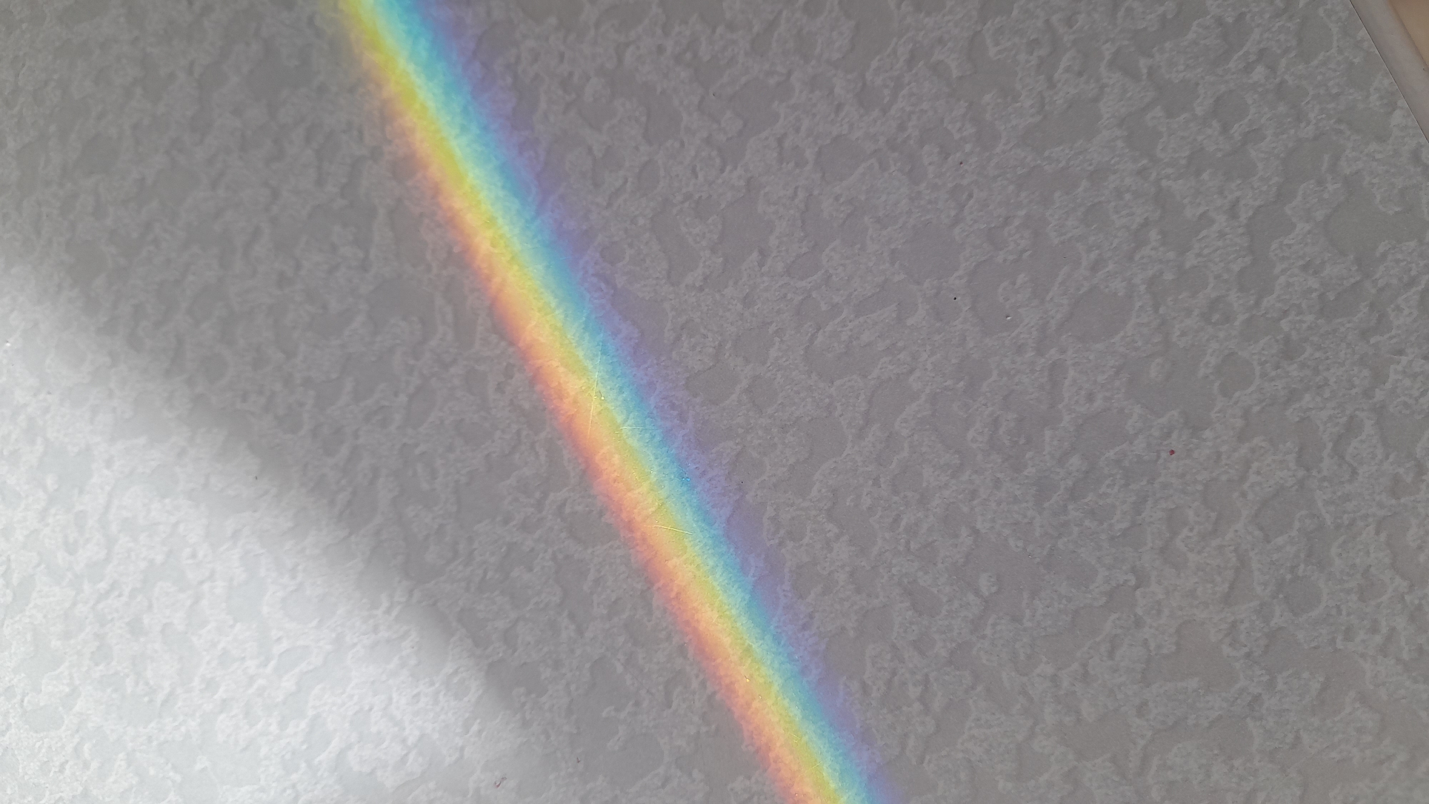 A rainbow is cast on a textured light gray surface. The rainbow spans from the upper right to the lower left corner of the image, with the full spectrum of colors visible. The background appears to be a wall or ceiling with an uneven texture. The location of the image is Saint Petersburg, Russia is not evident.