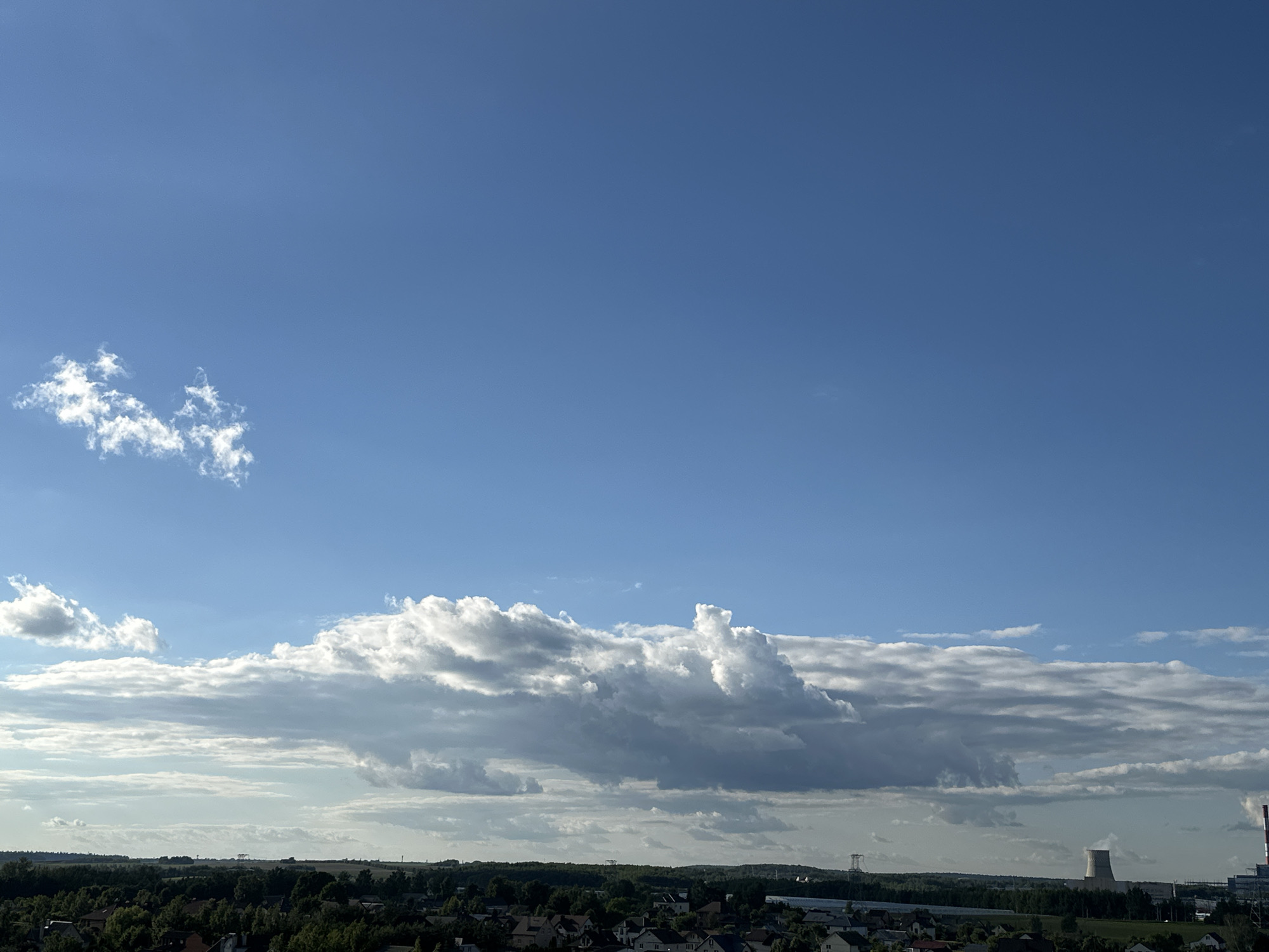 An aerial, eye-level view of a broad expanse of blue sky is captured above a horizontal line of trees and buildings. A few wispy white clouds are scattered across the upper portion of the frame, with a denser, larger formation of cumulonimbus clouds stretching across the mid-ground. These clouds vary in shade from bright white where illuminated by the sun to dark gray in their shadowed undersides, creating a dramatic contrast. The landscape below consists of a dense forest of green trees, interspersed with a settlement of houses. In the distance, on the right side of the frame, a large, cylindrical cooling tower of a power plant is visible, along with other industrial structures and transmission towers. The scene appears to be a suburban or rural area within the Шчомысліцкі сельскі Савет, Belarus, under a partly cloudy sky.