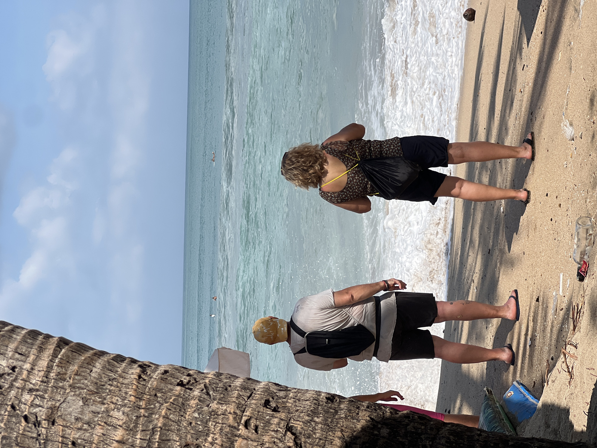 Two people, a man and a woman, stand on a sandy beach in Kuta, Indonesia, facing the ocean. The man, on the left, wears a light-colored t-shirt, dark shorts, a yellow baseball cap, and a black backpack. The woman, on the right, wears a patterned top and dark shorts, carrying a black bag over her shoulder. Gentle waves with white foam break on the shore. In the distance, a few individuals are visible in the turquoise water. The foreground of the beach shows scattered debris, including a coconut and a bottle. The scene is framed by the rough bark of a palm tree on the left side of the image.
