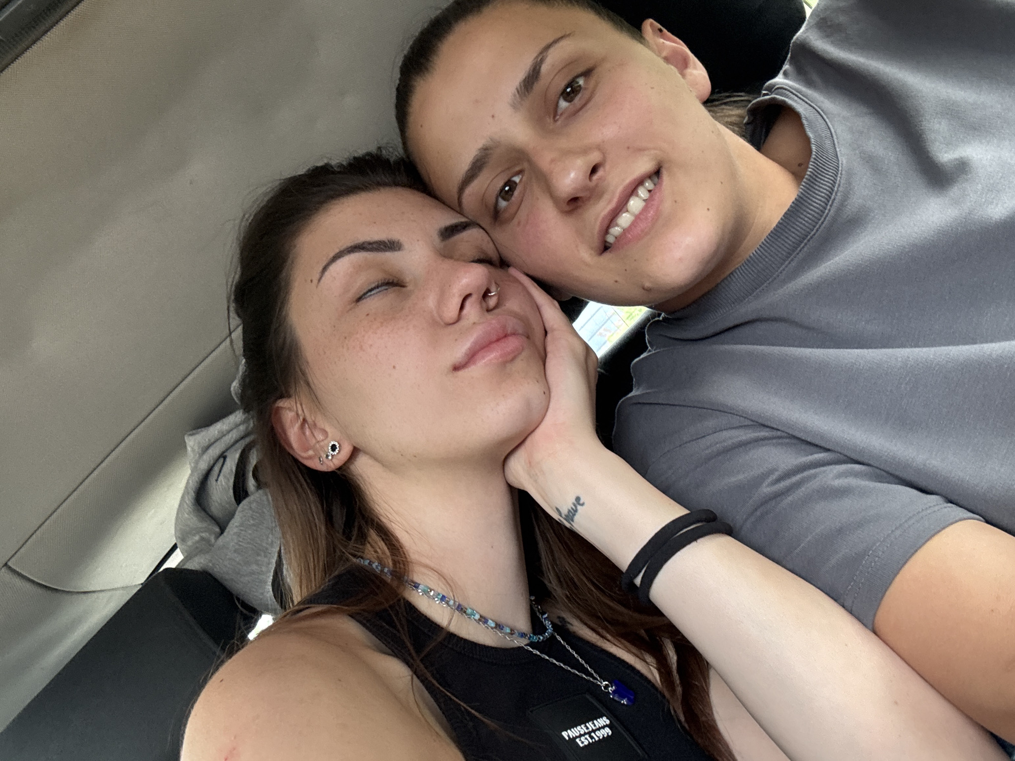 A close-up selfie shows two young women inside what appears to be a car in Dragoman, Bulgaria, during daytime. The woman on the left, with closed eyes, has long dark hair, freckles, a septum piercing, and multiple ear piercings. She rests her left hand on her jawline, revealing a small tattoo on her wrist that reads "have". She wears a black tank top with a white patch visible on her chest, bearing the text "PAUSE JEANS EST.1999", and a beaded necklace with a prominent blue pendant.
The woman on the right, with dark hair pulled back, has her eyes open and is looking towards the camera with a gentle smile, showing her white teeth. She is wearing a grey crew-neck t-shirt and has an ear piercing. The two are positioned intimately, with the woman on the left leaning into the other. The car's light-colored headliner and parts of the seat are visible in the background. The overall atmosphere suggests closeness and relaxation.