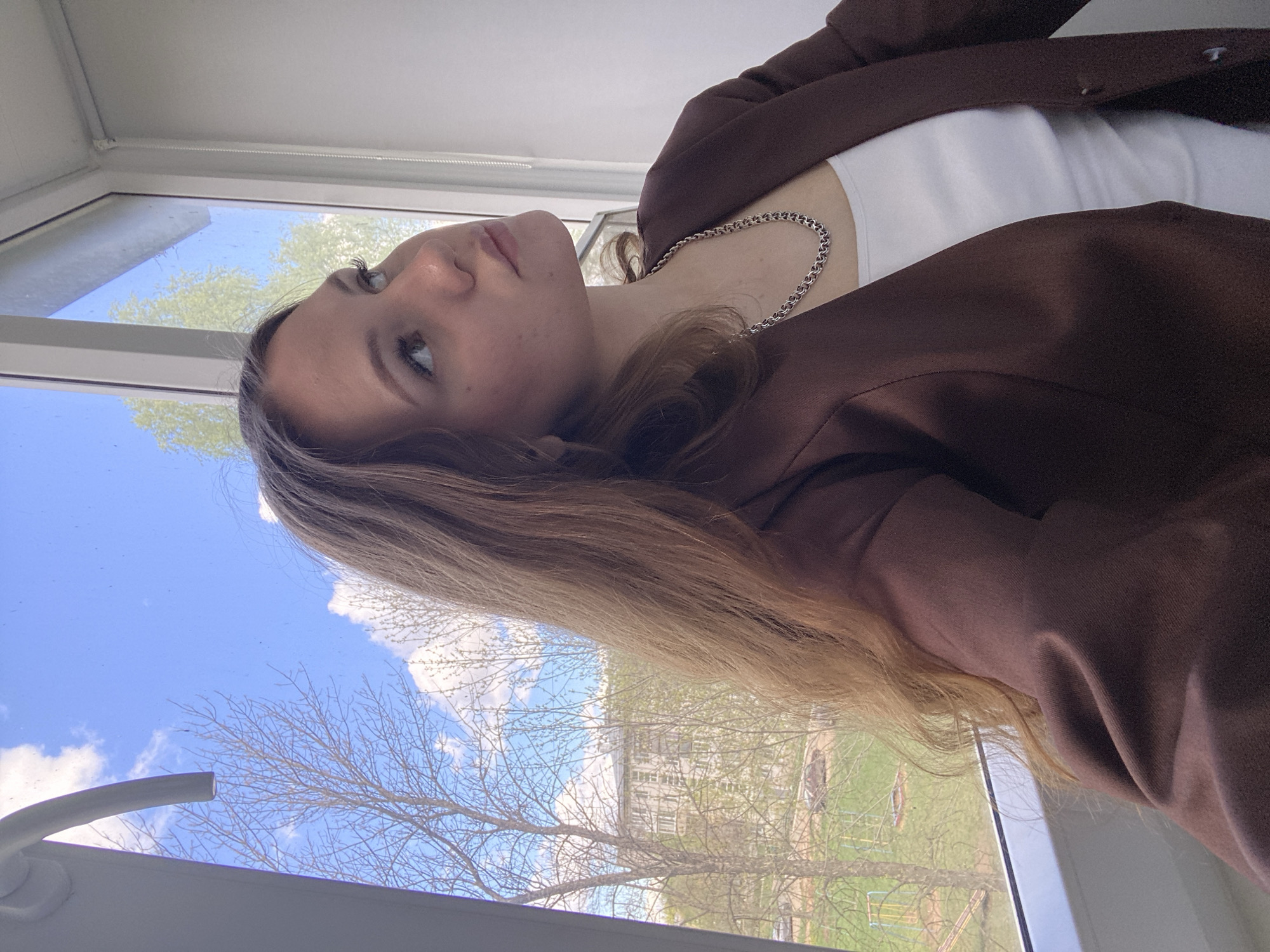 A young woman with long, light brown hair is sitting or standing by a window. She is wearing a white top and a brown jacket, and has a silver chain necklace. Outside the window, the sky is blue with white clouds, and a tree is visible in the foreground. In the background, there are buildings and a green lawn. It is not possible to determine the exact location within Izhevsk based on the information provided in the image.