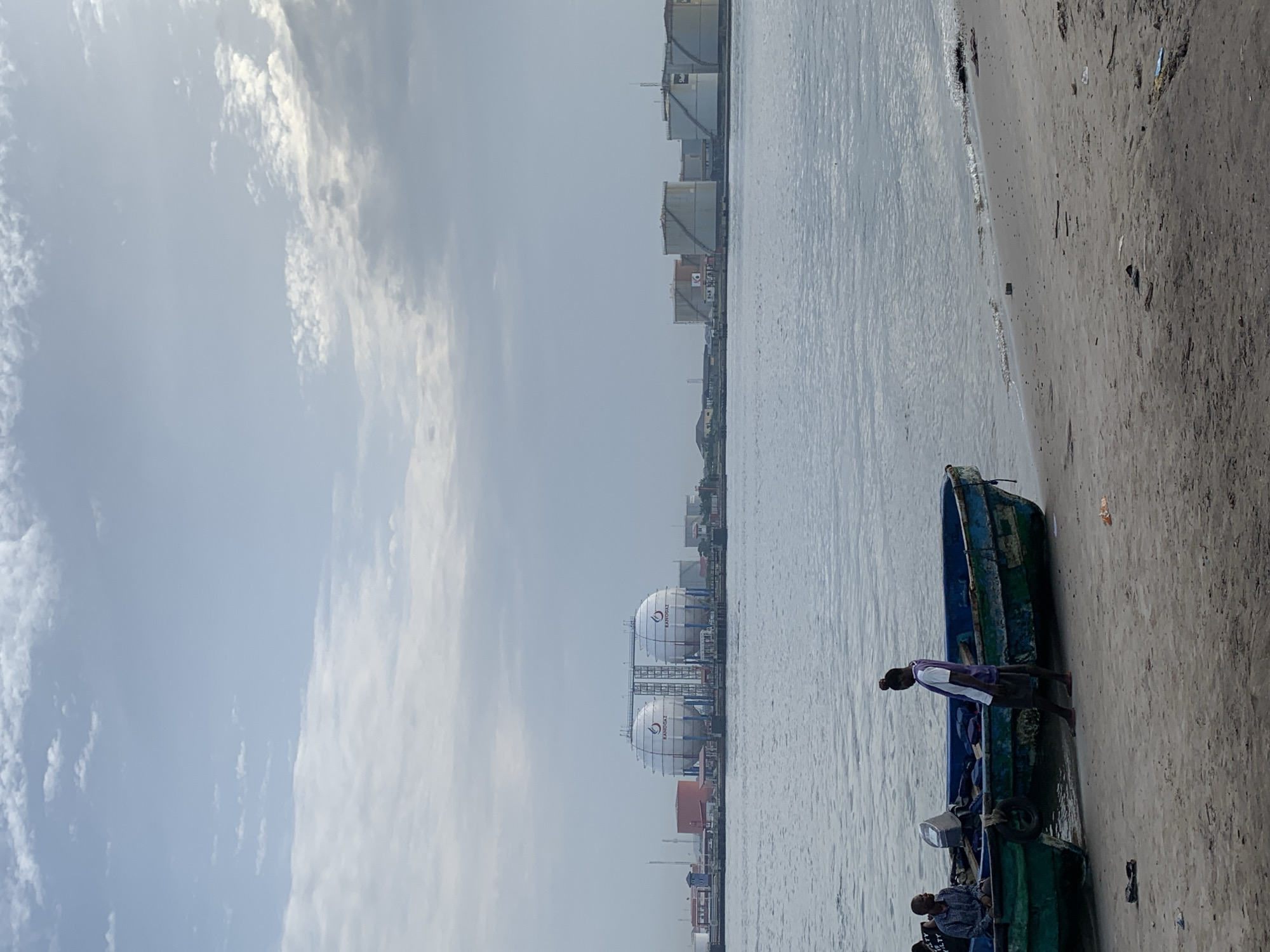 This image depicts a calm daytime scene along a waterfront in Ibasa, Nigeria, featuring both natural elements and significant industrial infrastructure.
In the foreground, a sandy or muddy bank stretches along the right side of the frame, where a small, dark blue and green boat is partially beached and partially in the water. A person, wearing a white shirt over a darker garment, stands prominently in this boat, facing towards the left. Other individuals are seated within the boat, their faces mostly obscured. More boats or parts of boats are visible adjacent to the main one.
A calm body of water, likely a river or bay, occupies the central and left-middle sections of the image, showing subtle ripples on its surface.
Across the water, the distant shoreline is lined with a series of large industrial structures. These include numerous cylindrical storage tanks and several large, prominent spherical tanks, all light in color. The text "RAPIDGAS" is clearly visible on many of these tanks, accompanied by a distinct logo that resembles a stylized number "6" or a similar symbol. On some of the rectangular structures, small symbols resembling Japanese flags and associated characters can be discerned. These facilities suggest a significant energy or gas storage and distribution operation.
The sky above is partly cloudy, with patches of bright white cumulus clouds set against a lighter blue or grey backdrop, indicating a bright, possibly overcast day. The scene conveys a sense of industrious activity alongside a natural waterway.