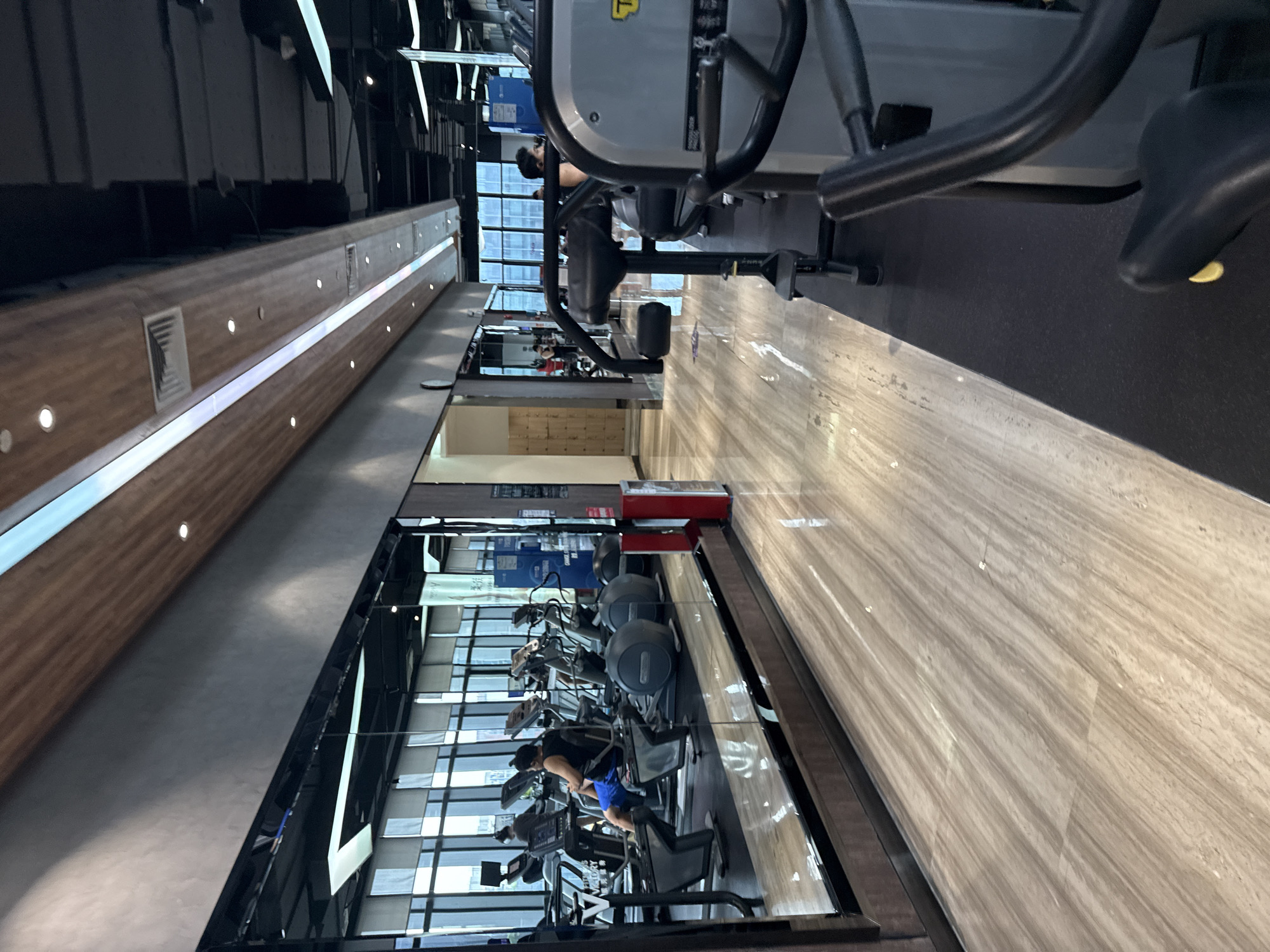 The image depicts an indoor gym located in the Haizhu District of China. In the foreground, a portion of an exercise machine with black handles and a black seat is visible. The floor is a glossy, light-colored wood-grain tile, reflecting the overhead lighting. To the left, a large mirrored wall reflects the gym's interior. This reflection shows several cardio machines, including treadmills and elliptical trainers, with people using them. One man in blue shorts and a black tank top is seated on an elliptical. Another person is visible on a treadmill in the reflection. The gym environment appears modern and well-lit, with a sleek ceiling featuring integrated lighting and wood paneling. In the background, more gym equipment and a large window with blinds are visible. A red trash receptacle is also present in the reflected area.