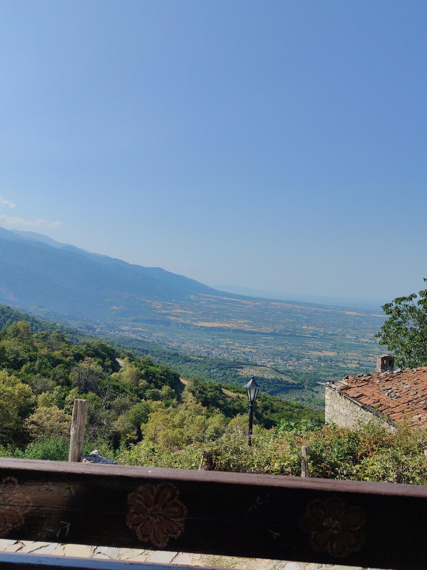 A vertical, medium-shot, outdoor photograph taken on a sunny day shows a scenic landscape view from an elevated vantage point in Palaios Panteleimonas, Greece. The foreground is occupied by a dark wooden railing with floral patterns. Beyond the railing, a stone building with a red tiled roof and a chimney is partially visible on the right. A black, traditional-style lamppost stands in front of the building. The middle ground features lush green trees and vegetation, with a winding road visible in the distance. The background is dominated by a wide valley with scattered houses and agricultural fields, leading to a range of hazy blue mountains that fade into the clear, bright blue sky.