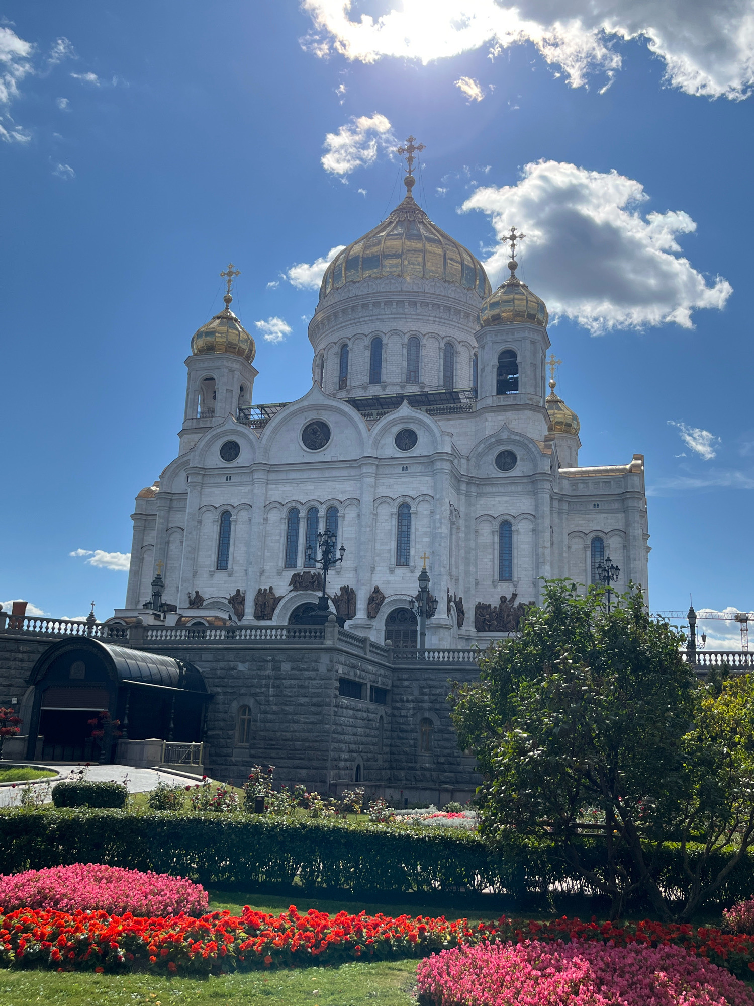 The image shows the Cathedral of Christ the Saviour in Moscow, Russia. The white stone cathedral features multiple golden domes and crosses, with a bright blue sky and scattered white clouds as the backdrop. In the foreground, there are manicured green lawns and vibrant flowerbeds filled with red and pink flowers. A lush green tree is situated to the right of the cathedral. The cathedral is elevated, with a stone-clad lower level and decorative sculptures adorning its facade. A pathway with black railings is visible leading to an arched entrance. The overall scene depicts a sunny day at a prominent landmark.