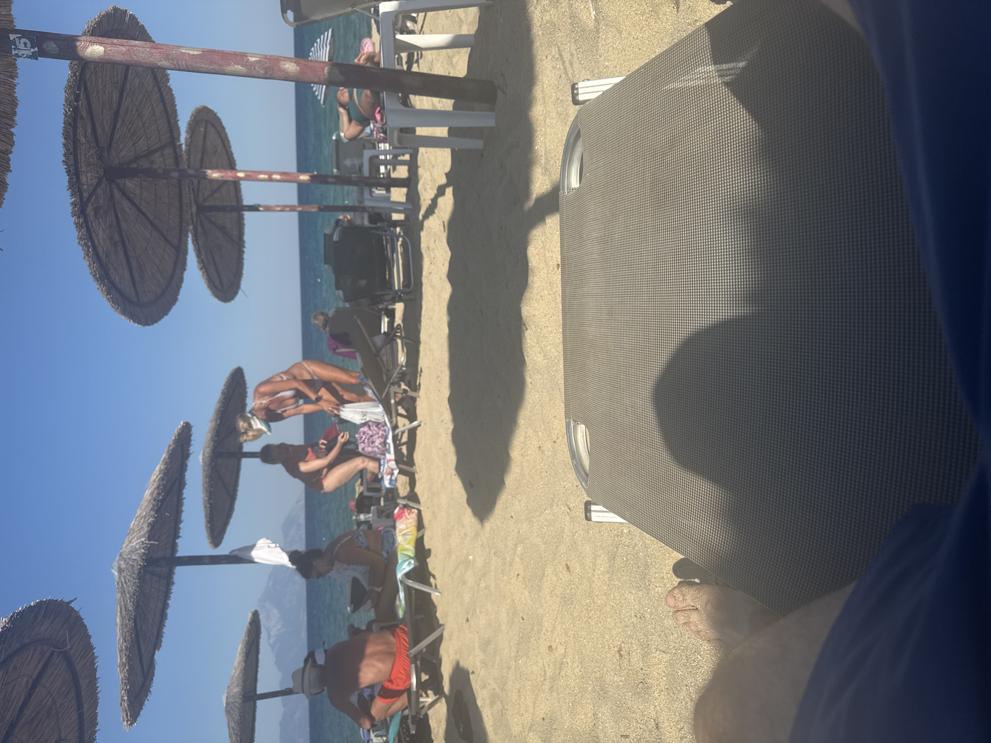 A first-person perspective shot of a beach in Toroni Municipal Unit, Greece, shows a sun lounger in the foreground with a person's legs and feet resting on it. In the midground, beach umbrellas provide shade for several people sitting on loungers. The sea is visible in the background, with mountains in the far distance. The sand is light brown and the sky is clear blue. The scene suggests a relaxed atmosphere on a sunny day.