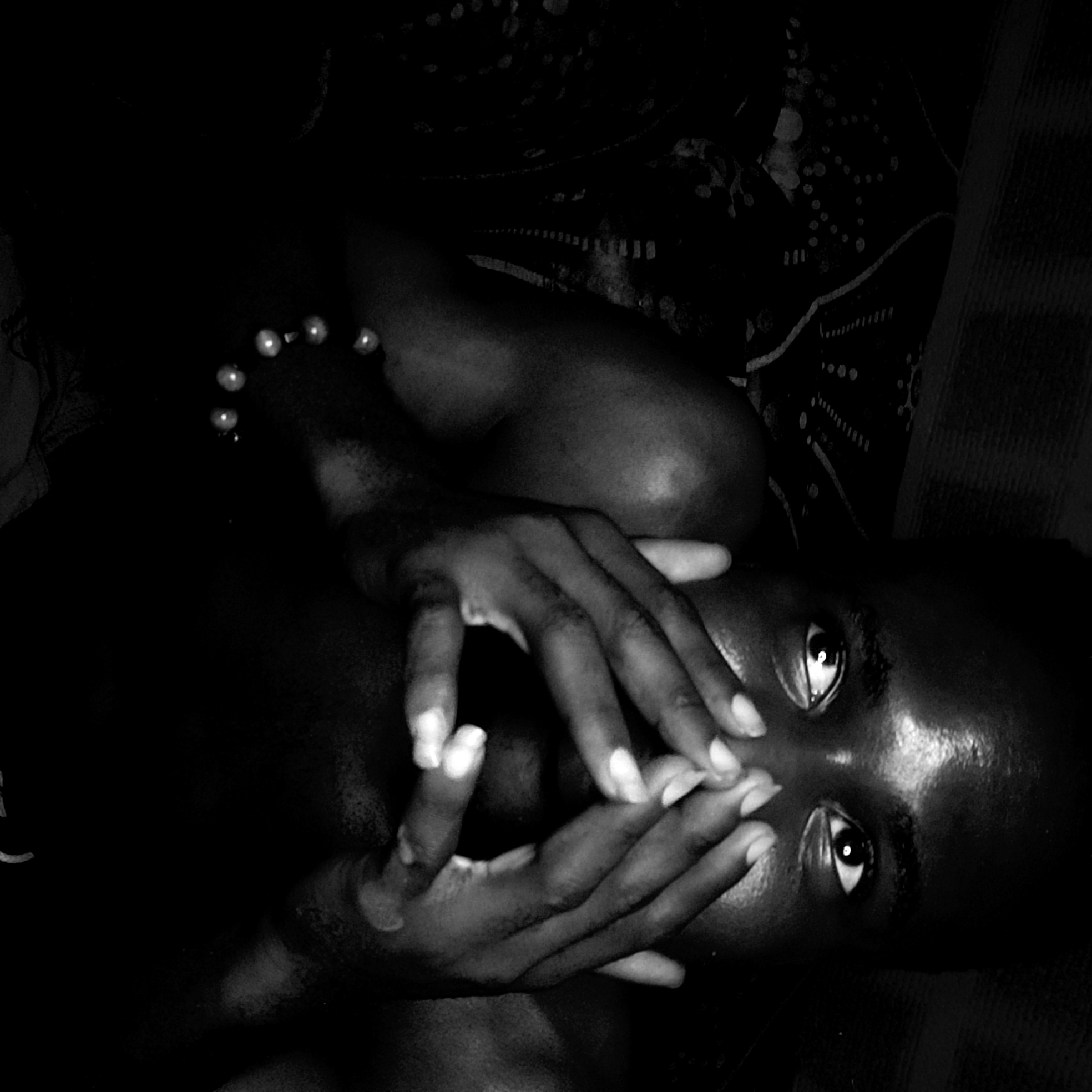 The image is a black and white, close-up shot of a dark-skinned person lying down. The person's face is visible with eyes open, and their hands are positioned over their mouth. A string of pearls is visible around the person's neck or arm. The background is dark and out of focus, suggesting a bed with patterned sheets and a wooden headboard. The image was taken at an indoor location in Narayi, Nigeria.