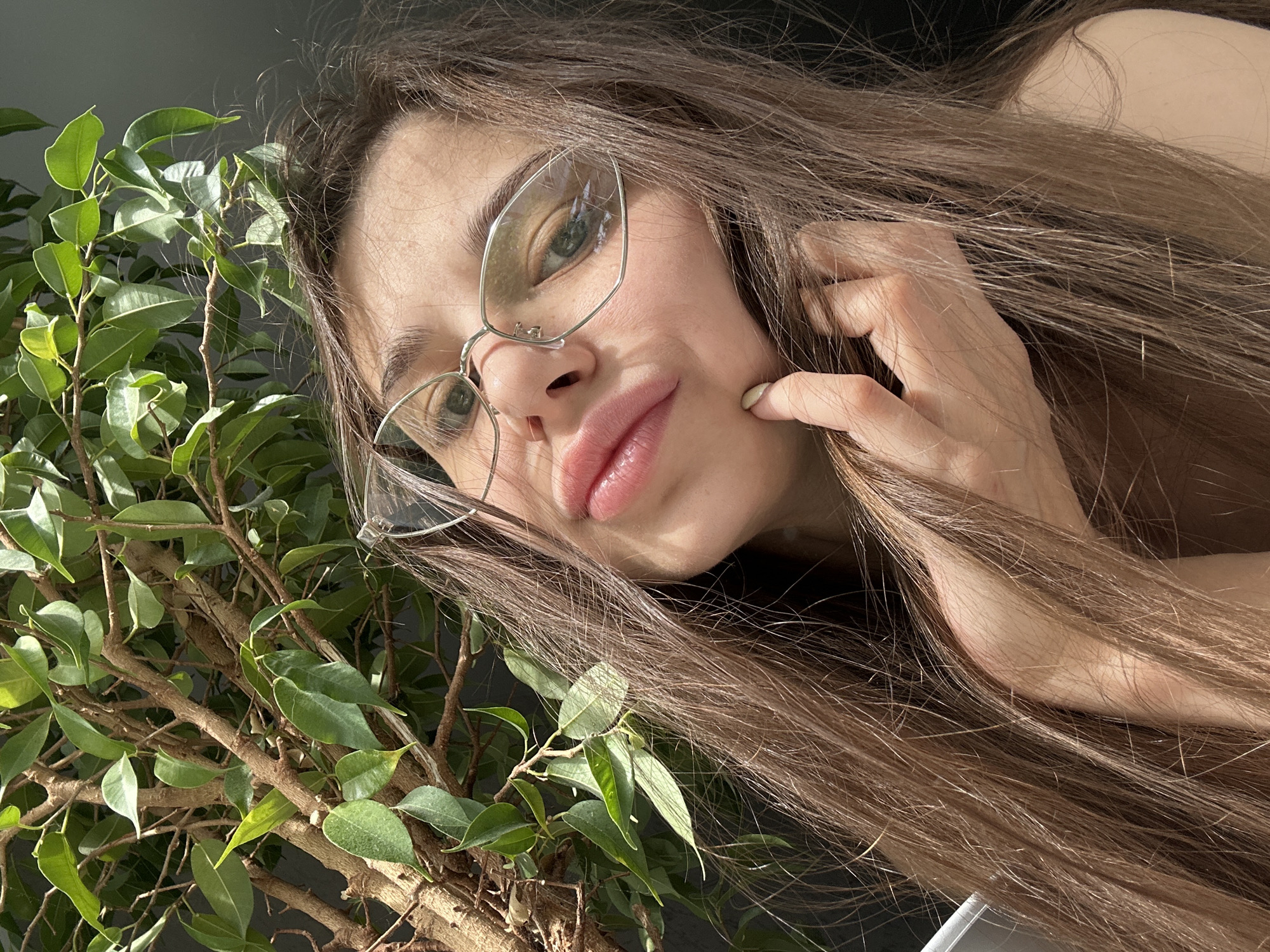 A woman with long brown hair and glasses is positioned to the right of a leafy plant. Her hand is near her cheek. The background is a neutral gray. There is no indication this image was taken in Izhevsk, Russia.