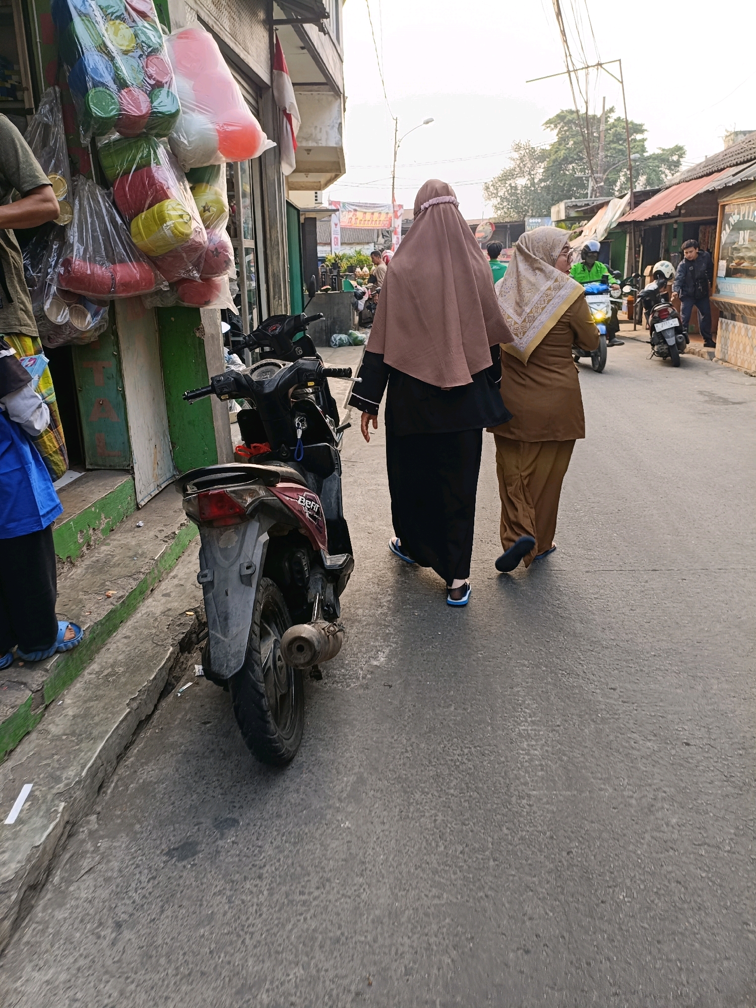 The image depicts a street scene in Tangerang, Indonesia, likely in a local market or commercial area. Two women are walking away from the camera, both wearing hijabs and modest clothing. The woman on the left wears a brown hijab and a black abaya, and the woman on the right wears a beige hijab and a brown outfit. They are walking on an asphalt street next to a row of shops. In the foreground, a red and black motorcycle is parked on the side of the road. Shops are visible on the left, with items like colorful plastic containers hanging from the storefront. Further down the street, other people and motorcycles are present, suggesting a busy urban environment. The overall atmosphere is that of a typical Indonesian street scene.