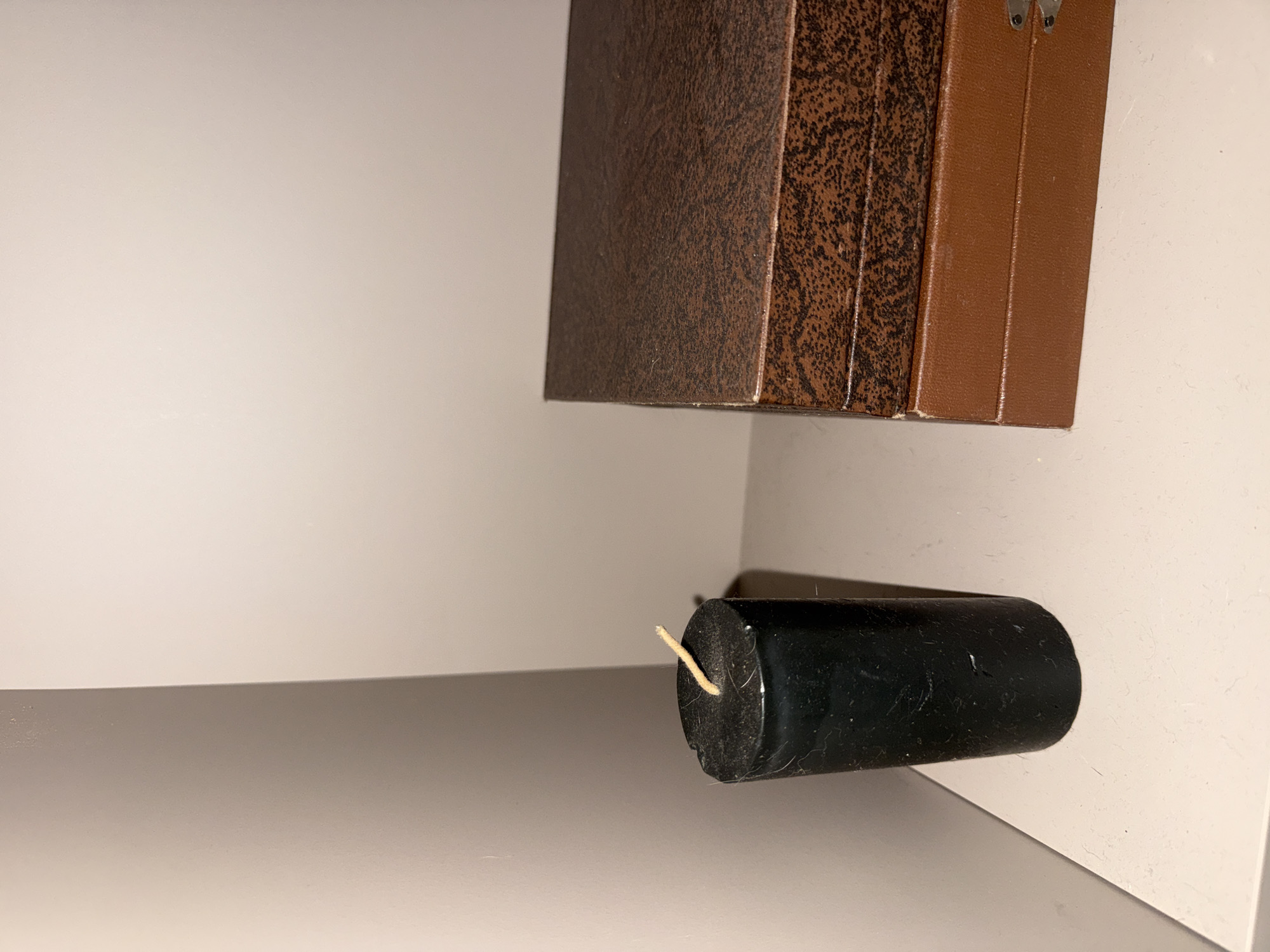 An indoor scene in Berezyna, Ukraine, depicts a still life arrangement. On a light-colored, flat surface, a cylindrical black candle stands upright on the left. The candle has a light-colored wick protruding from its top and exhibits minor surface imperfections, possibly including dust or lint. To the right of the candle, two rectangular boxes are stacked. The top box is dark brown with a textured, mottled pattern. The bottom box is a solid, lighter reddish-brown color with a subtly ribbed surface, and a small silver-colored metal clasp is visible on its right side. The background consists of a plain, light-colored wall, forming an interior corner, suggesting the items are placed against a wall or in a niche. There are no people present, and no actions or interactions are depicted.