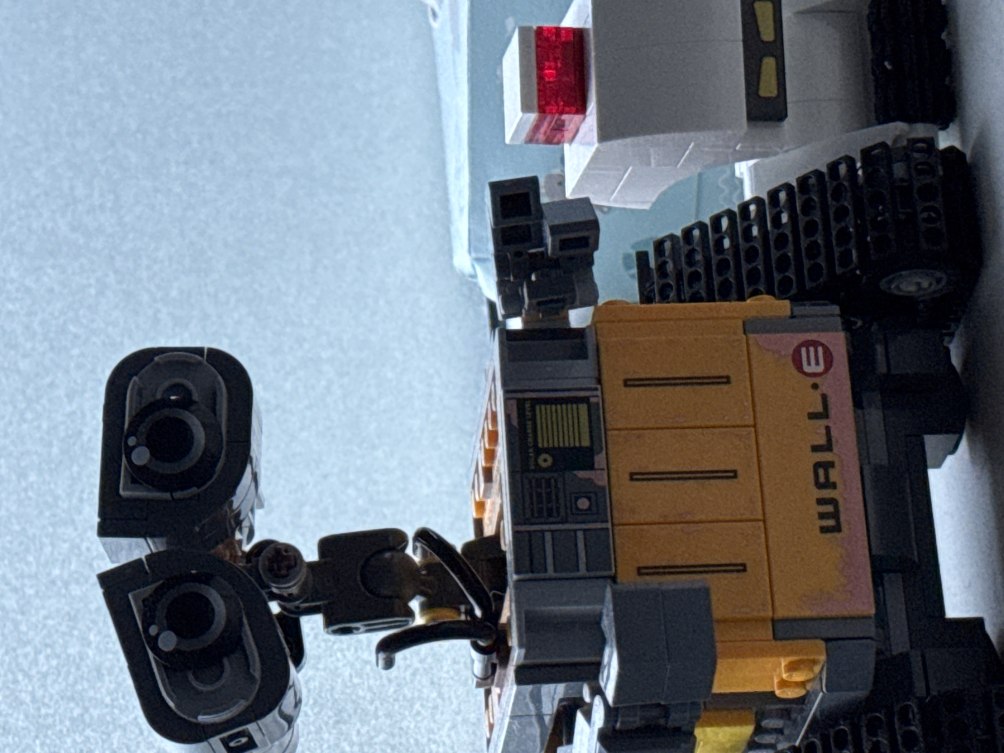 The image displays two stationary LEGO models against a light blue, out-of-focus background. The primary subject, a model of the robot WALL-E, occupies the left and central foreground. Its head features two black, rectangular "eyes" with circular lenses and white highlights, connected by an articulated dark grey structure. The robot's body is predominantly orange and grey, displaying the text "WALL•E" and an 'E' logo in black on its lower front panel. A faint pinkish discoloration is visible on the orange bricks below the text. The top section includes printed details such as a "SOLAR CHARGE LEVEL" indicator and orange studs. Black segmented tracks are visible on the left side of the WALL-E model, resting on a textured light grey surface. To the right, a partially visible white LEGO model, possibly a second robot, is present, topped with a small, transparent red brick capped with white. The background lacks specific features, preventing geographical identification. No people are present. The location of Riga, Latvia, cannot be determined from the visual information.